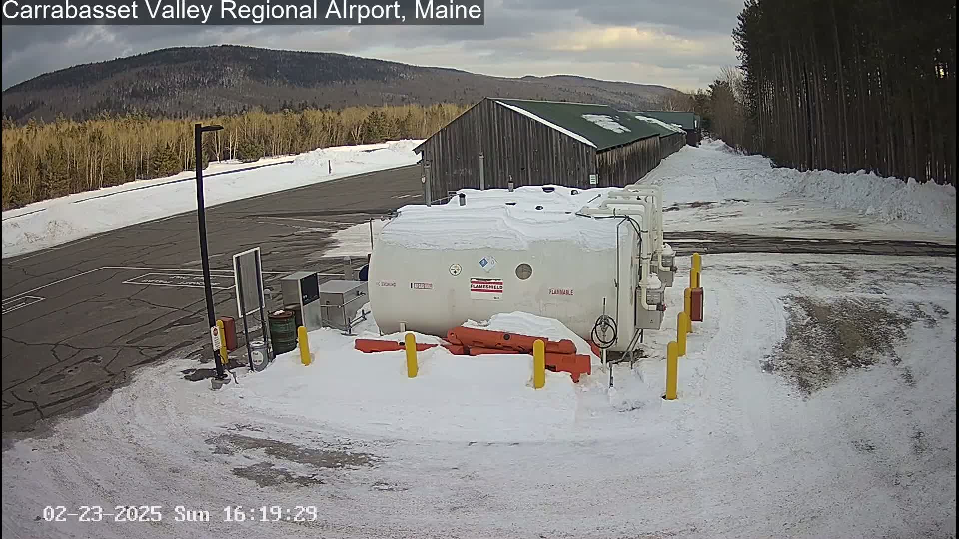 A snow-covered airport tarmac with a large white fuel tank, a long wooden building, and a background of snow-covered trees and mountains under an overcast sky.