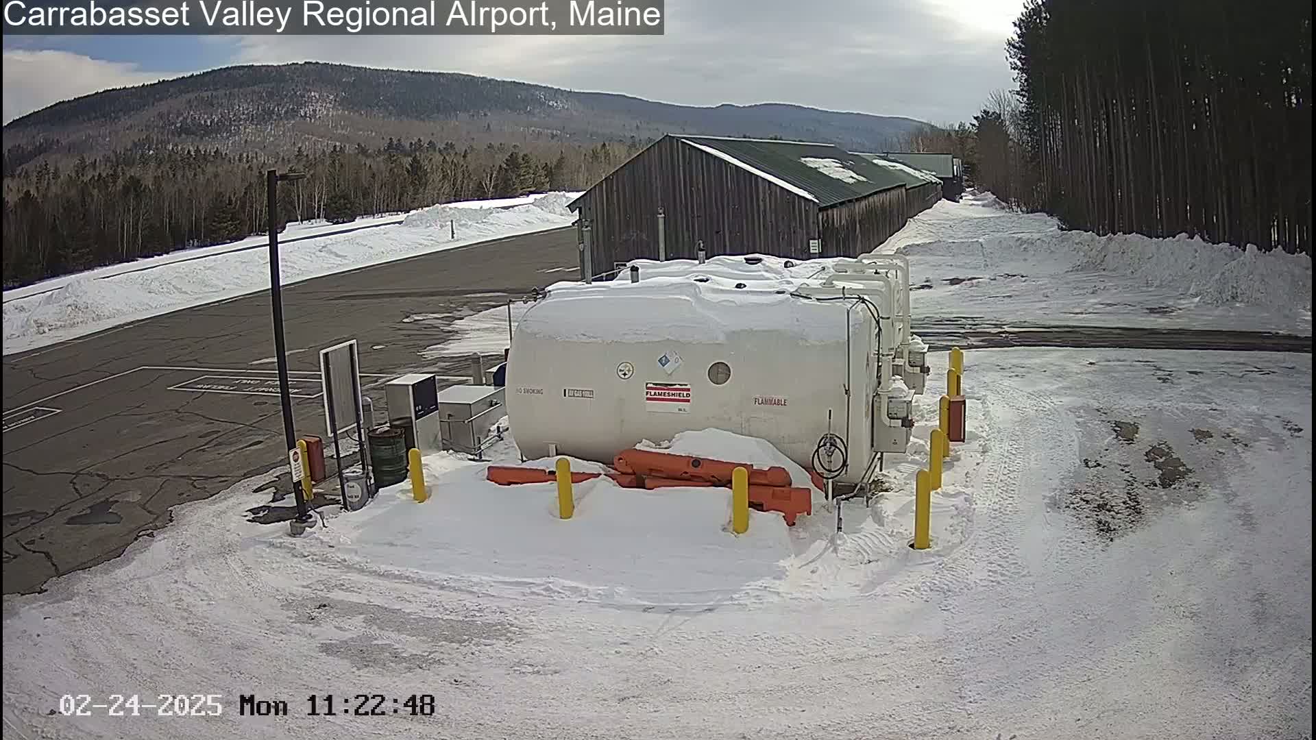 A snow-covered airport tarmac with a large white fuel tank, a long building, and a mountain range in the background under a partly sunny sky.