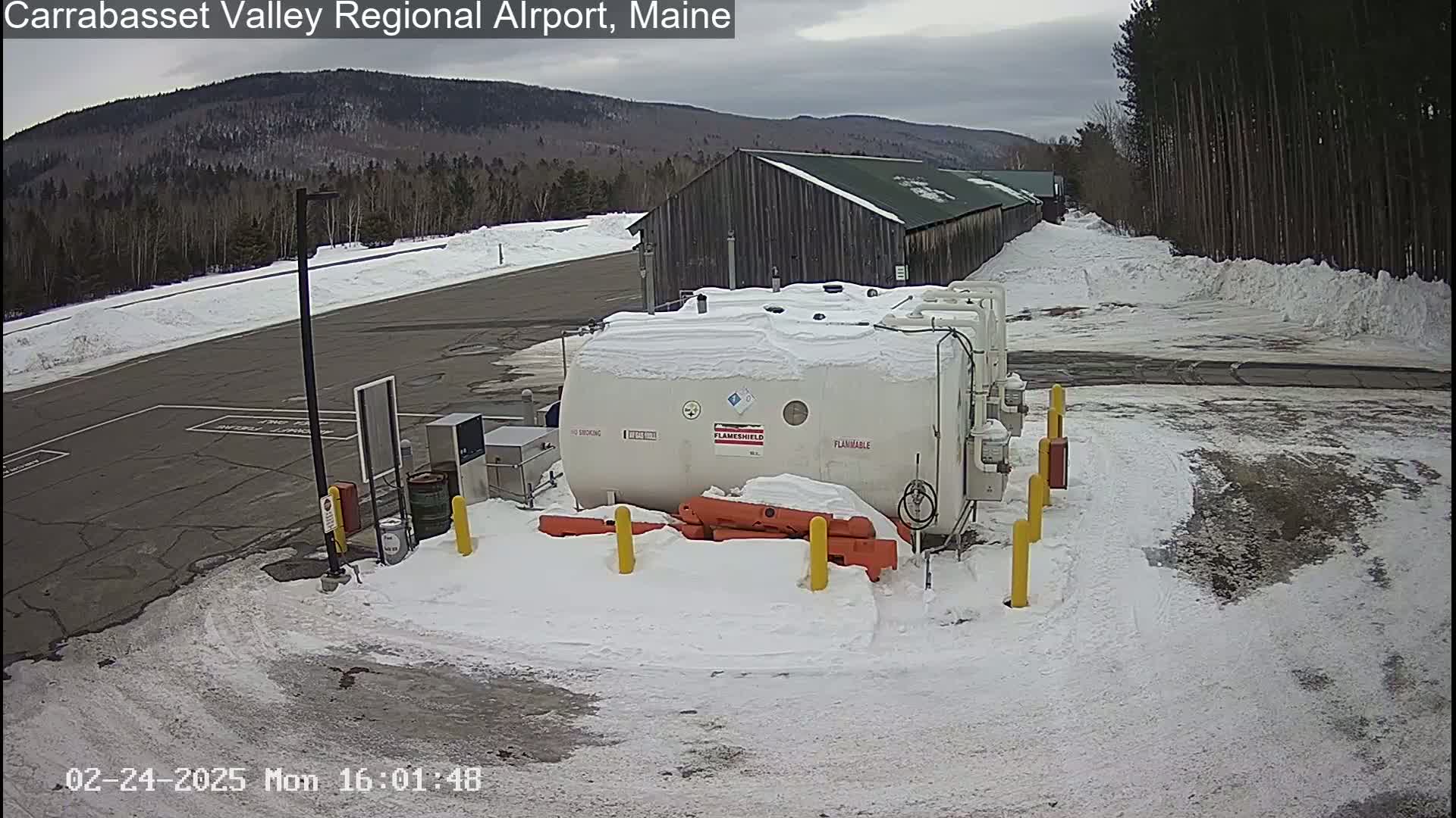 A snow-covered airport tarmac with a large white tank, a long building, and a snowy landscape in the background under a cloudy sky.