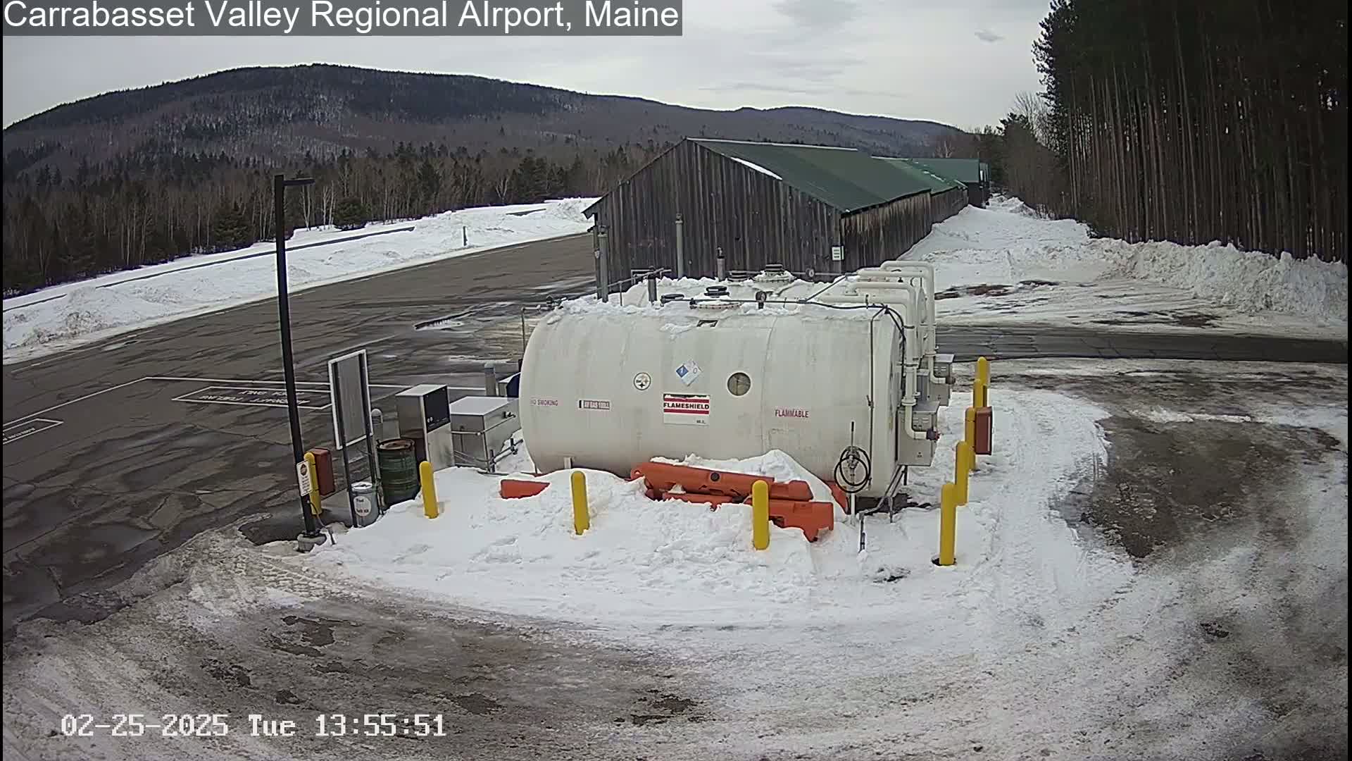 A large white tank sits in a snowy airport parking lot next to a long wooden building, with a mountain range visible in the background.