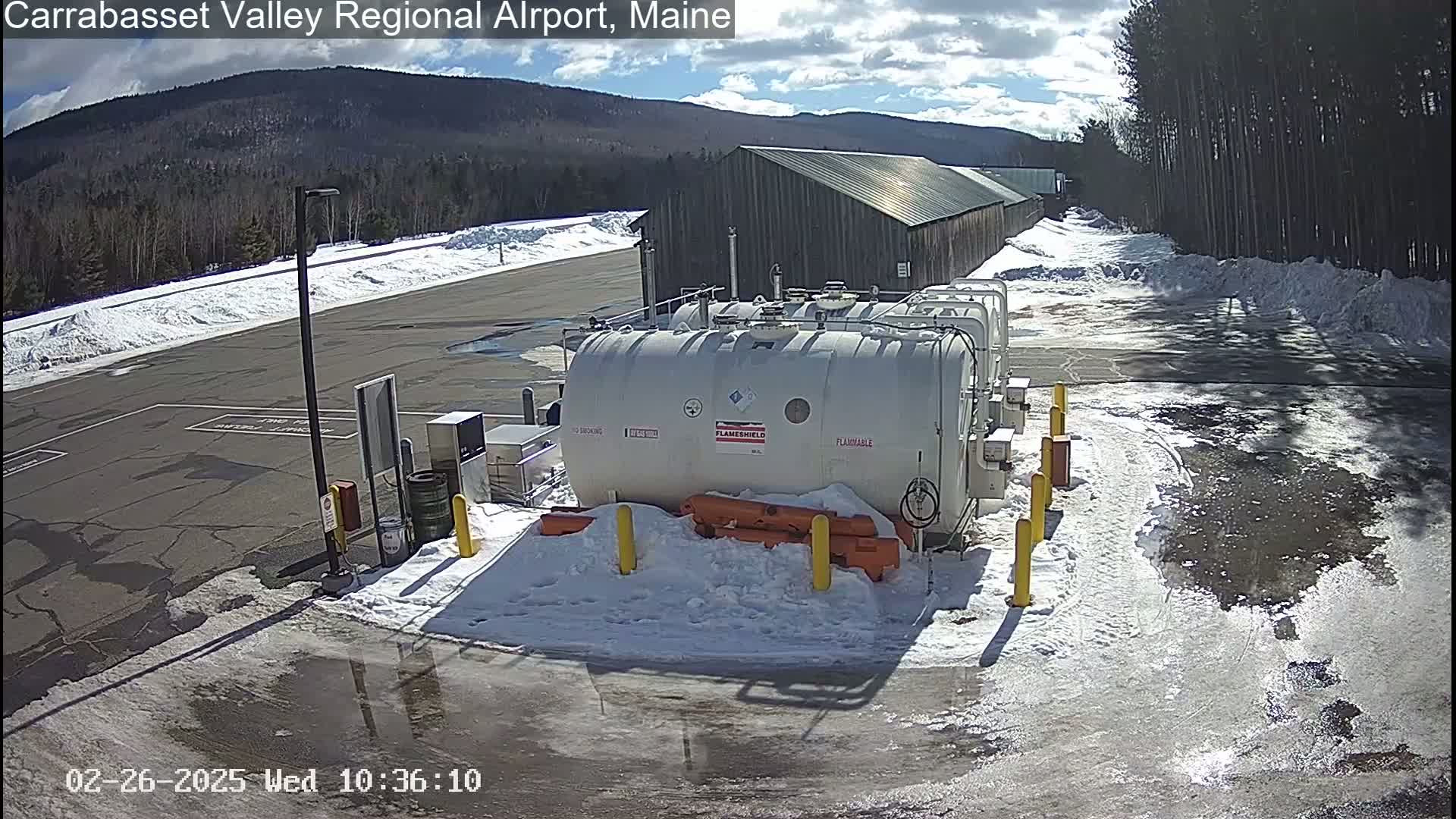 Several large white fuel tanks sit in a snowy airport parking lot under a partly sunny sky, with a long wooden building and distant mountains visible in the background.