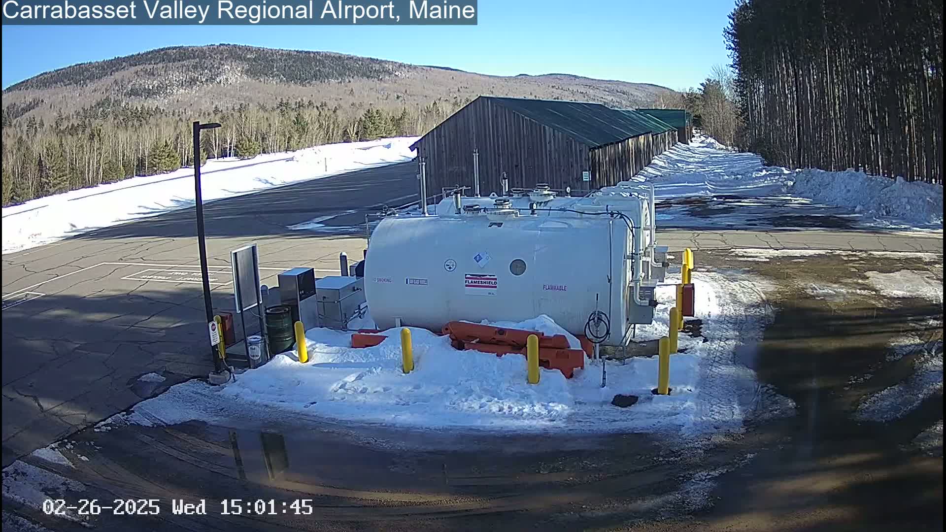 A large, white fuel tank sits in a snowy airport parking lot on a sunny day, with a long, dark building and a line of trees in the background.