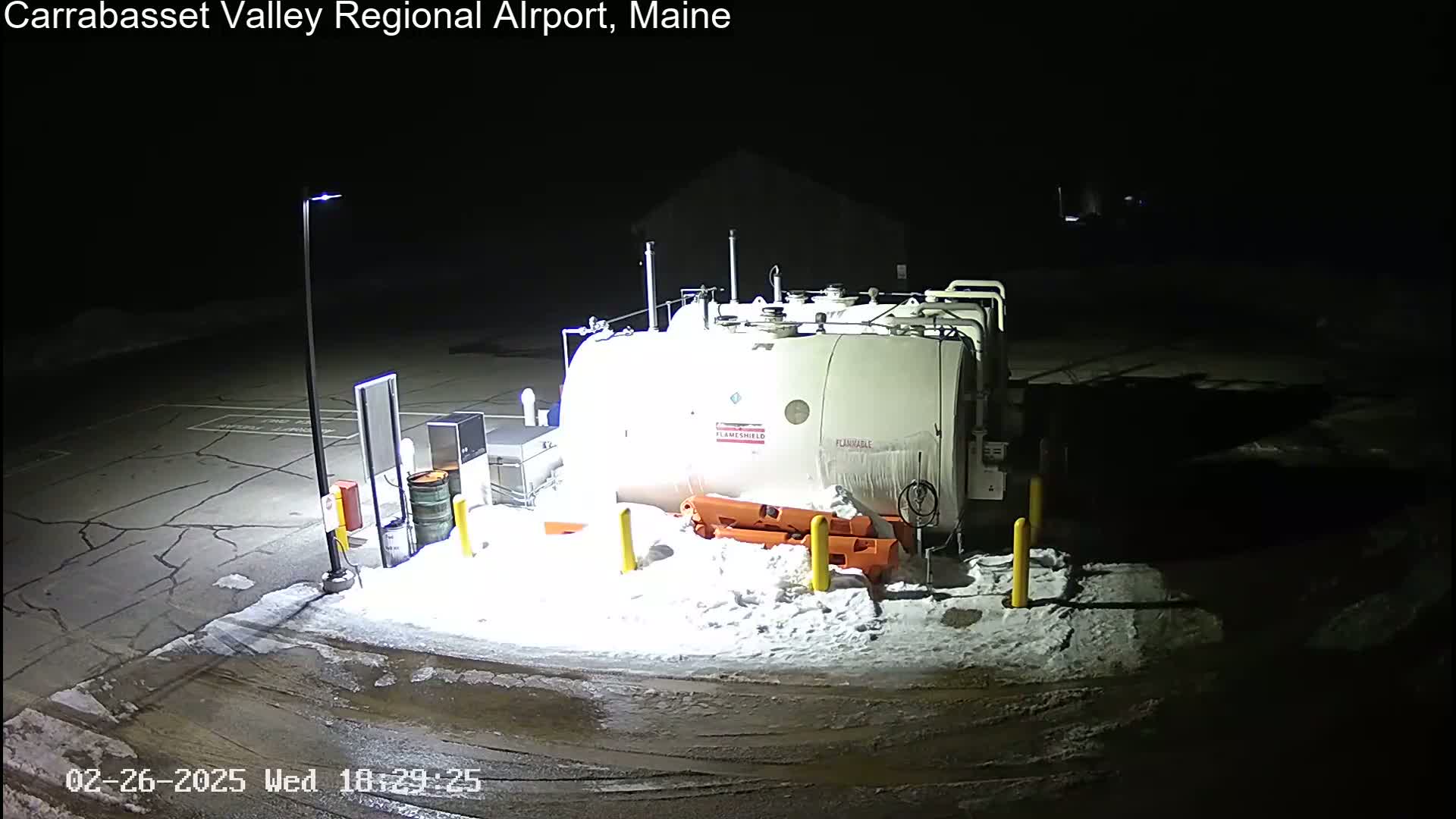 A large, white fuel tank sits in a snow-covered airport parking lot at night, illuminated by artificial lights.