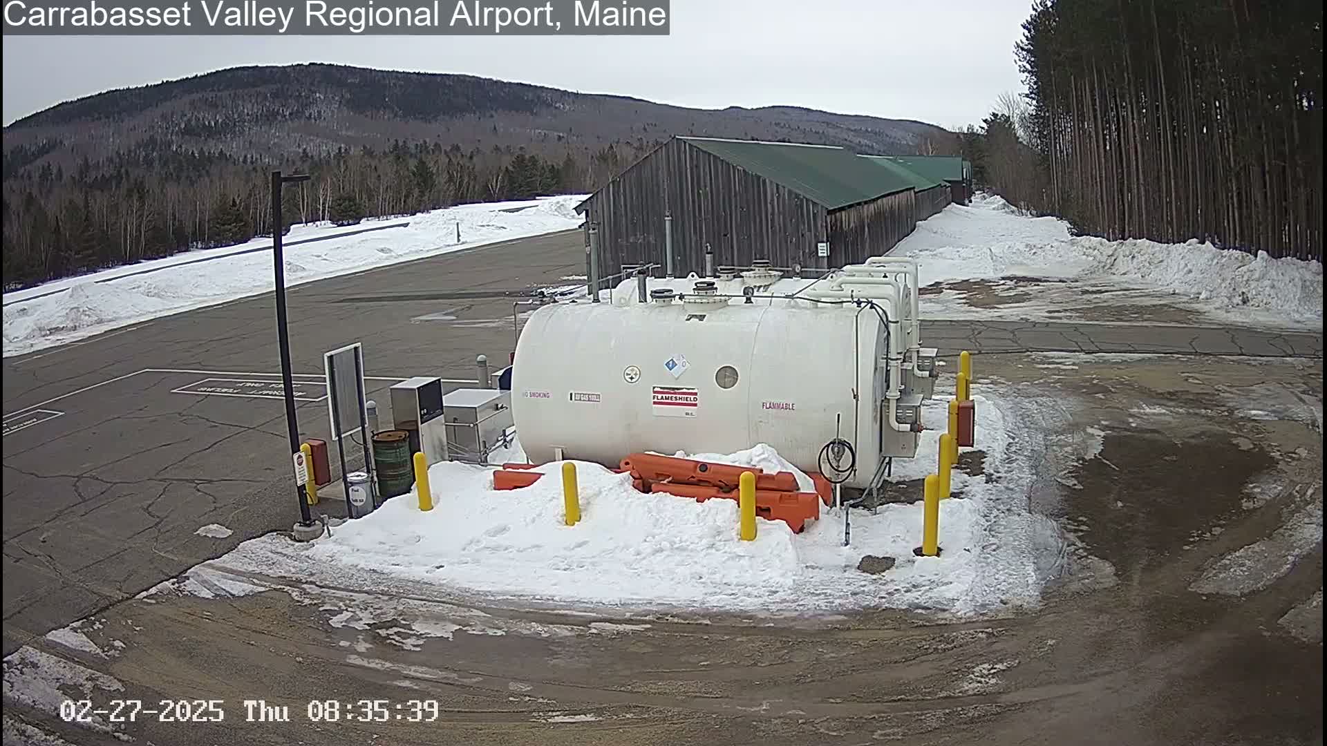 A large white fuel tank sits in a snow-covered airport parking lot, with a building and a wooded hillside in the background under an overcast sky.