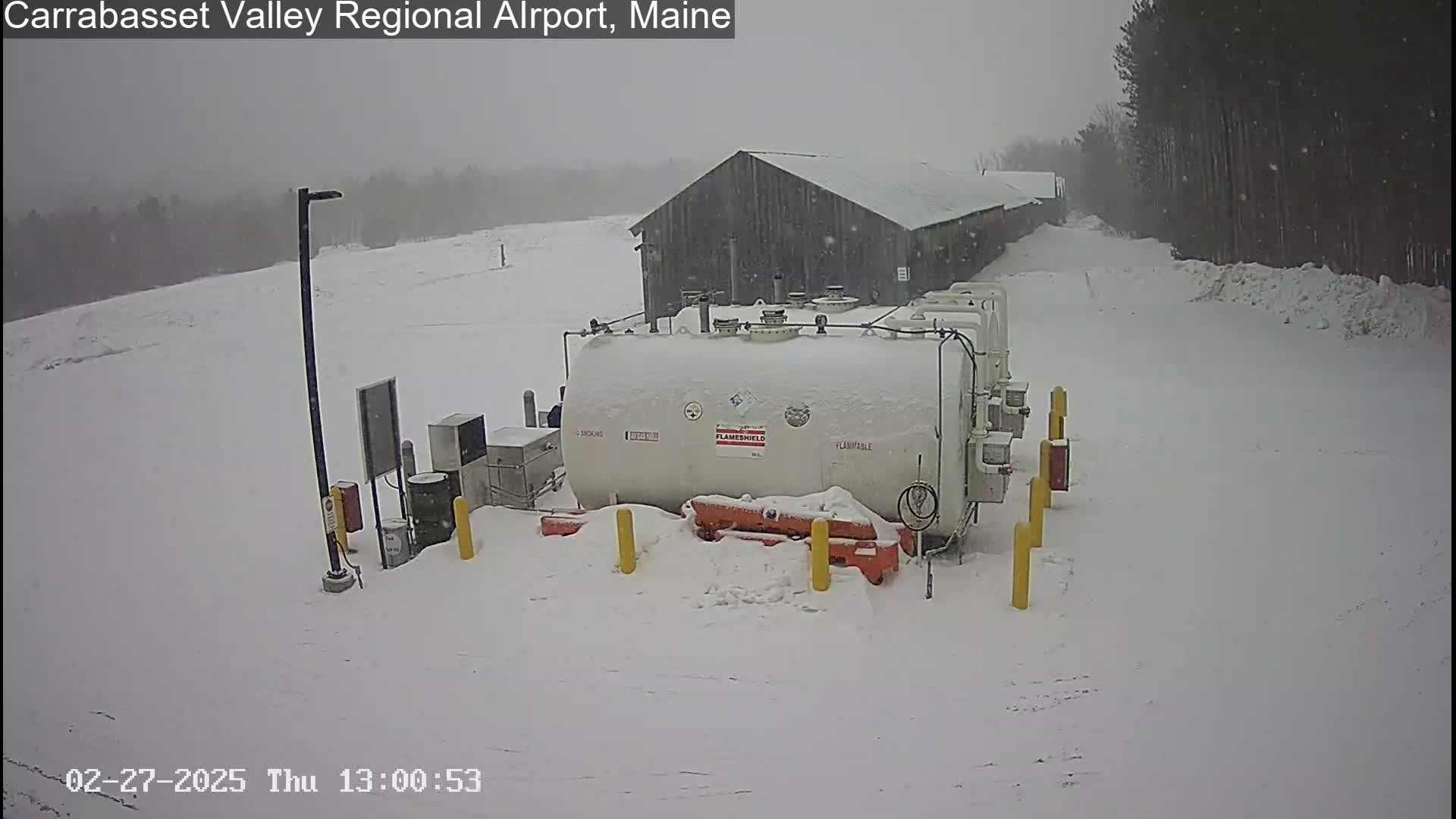 A large white fuel tank sits in the snow next to a dark-colored building during a snowfall.