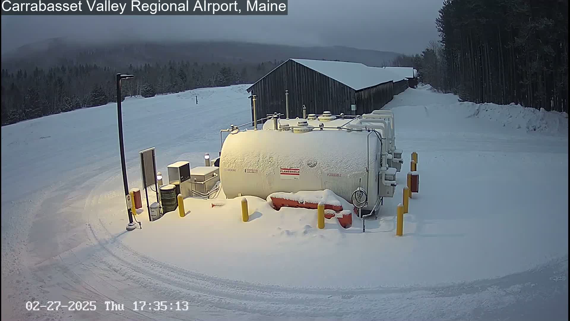 A snow-covered fuel tank sits in a snowy area near a long, snow-covered building, under a gray sky.