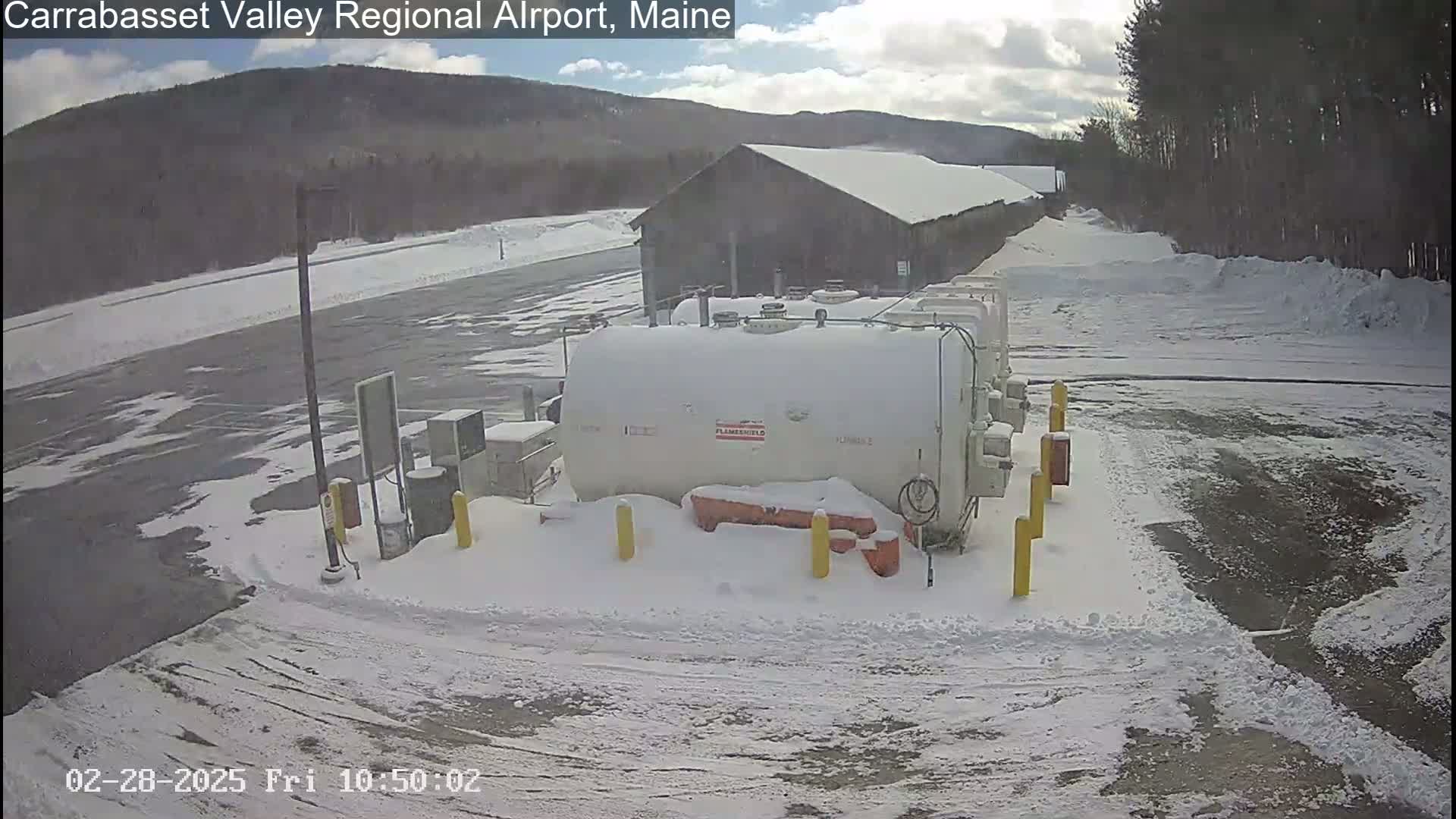 A snow-covered fuel depot sits near a building in a snowy landscape, with a mountain visible in the background under a partly cloudy sky.