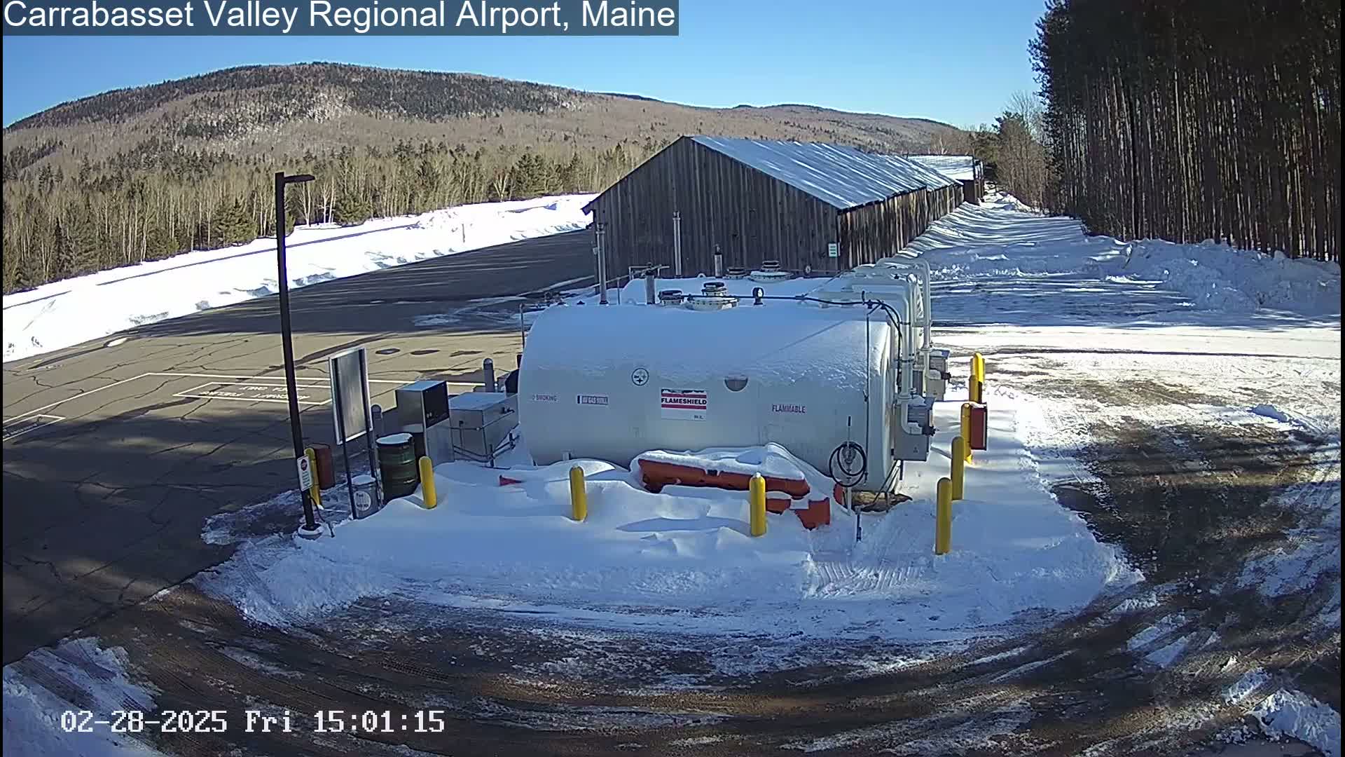 A snow-covered fuel storage area sits in front of a long wooden building, with a sunny, snow-covered landscape and distant mountains in the background.