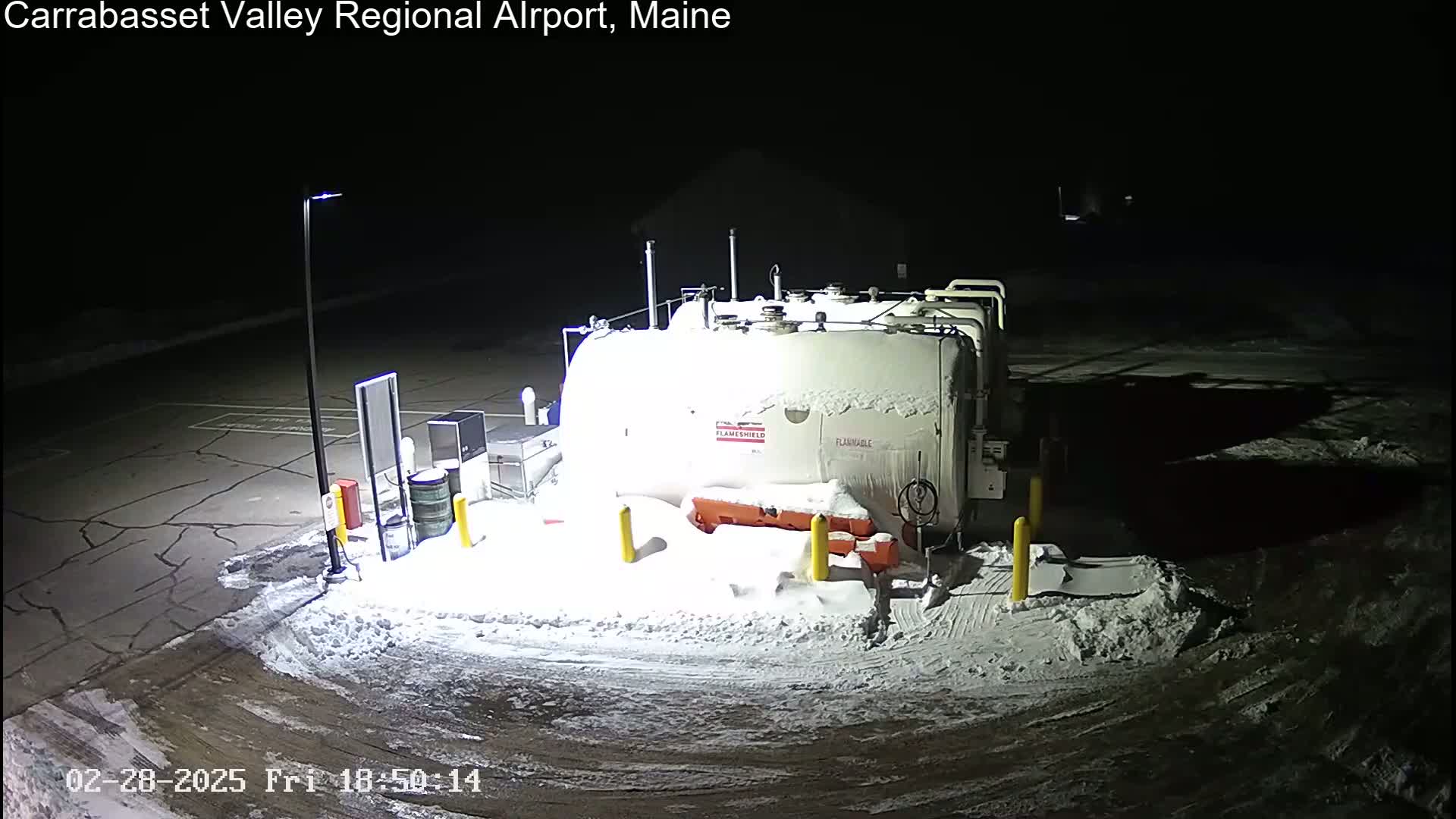 A snow-covered fuel tank sits in a mostly empty, snow-covered lot at night, illuminated by a single lamppost.