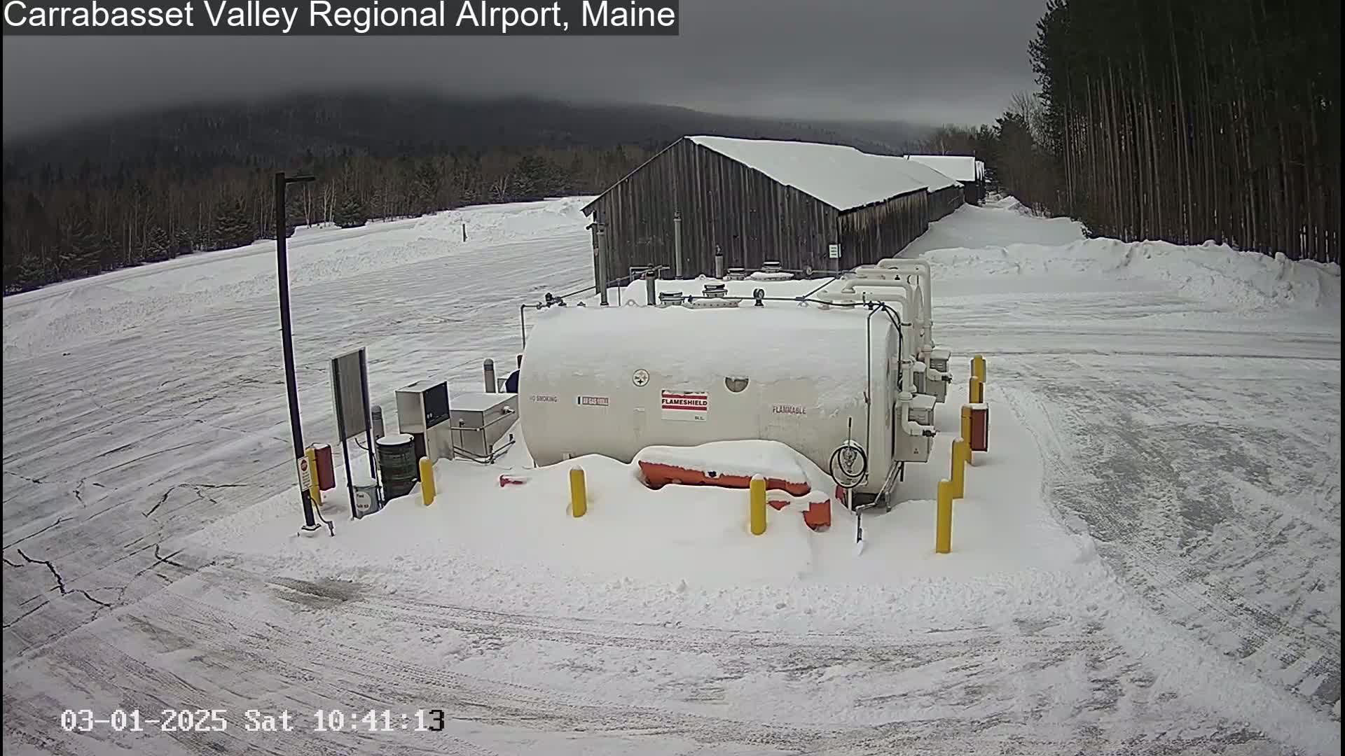 A snow-covered airport fuel storage area with a large white tank, a dark-colored building, and a mostly snow-covered tarmac under an overcast sky.