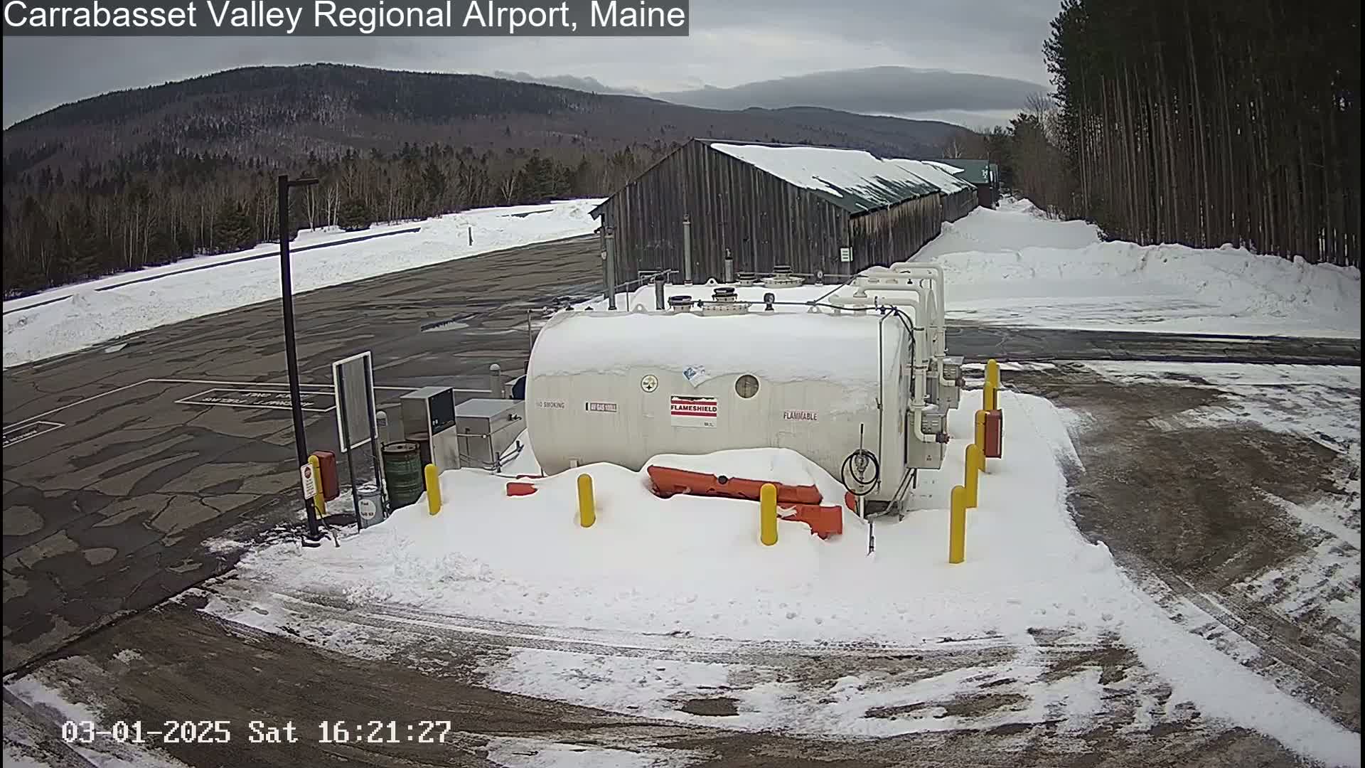 A snow-covered airport fuel storage area with a large, white fuel tank, a nearby building, and a mountainous background under an overcast sky.