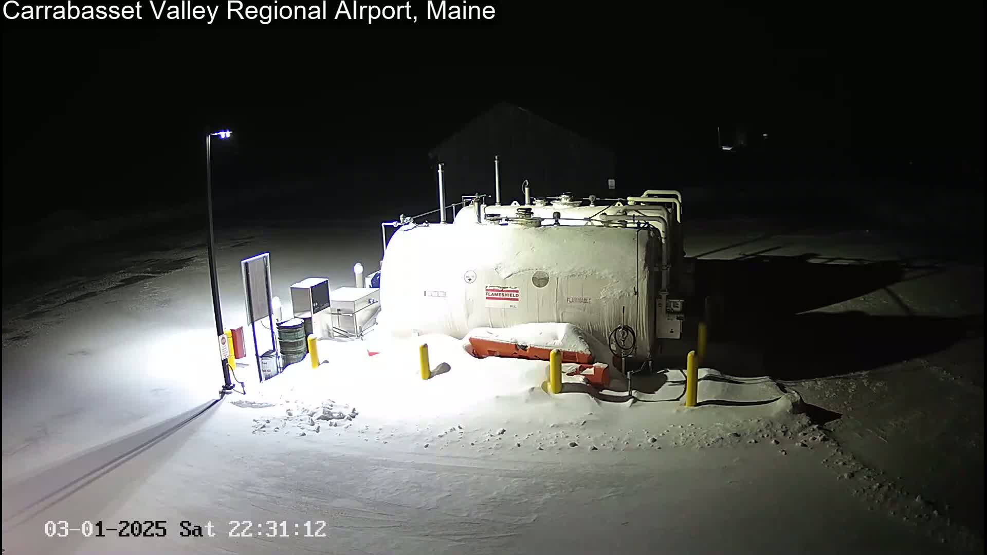 A large, snow-covered fuel tank sits in a snow-covered area at night, illuminated by a single lamppost.