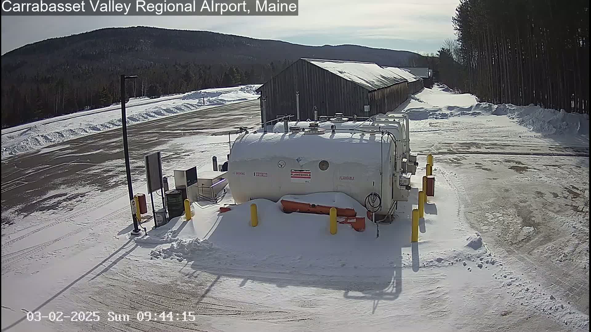 A snowy airfield with several large white tanks, a long shed-like building, and a partly snow-covered parking lot under a sunny sky.
