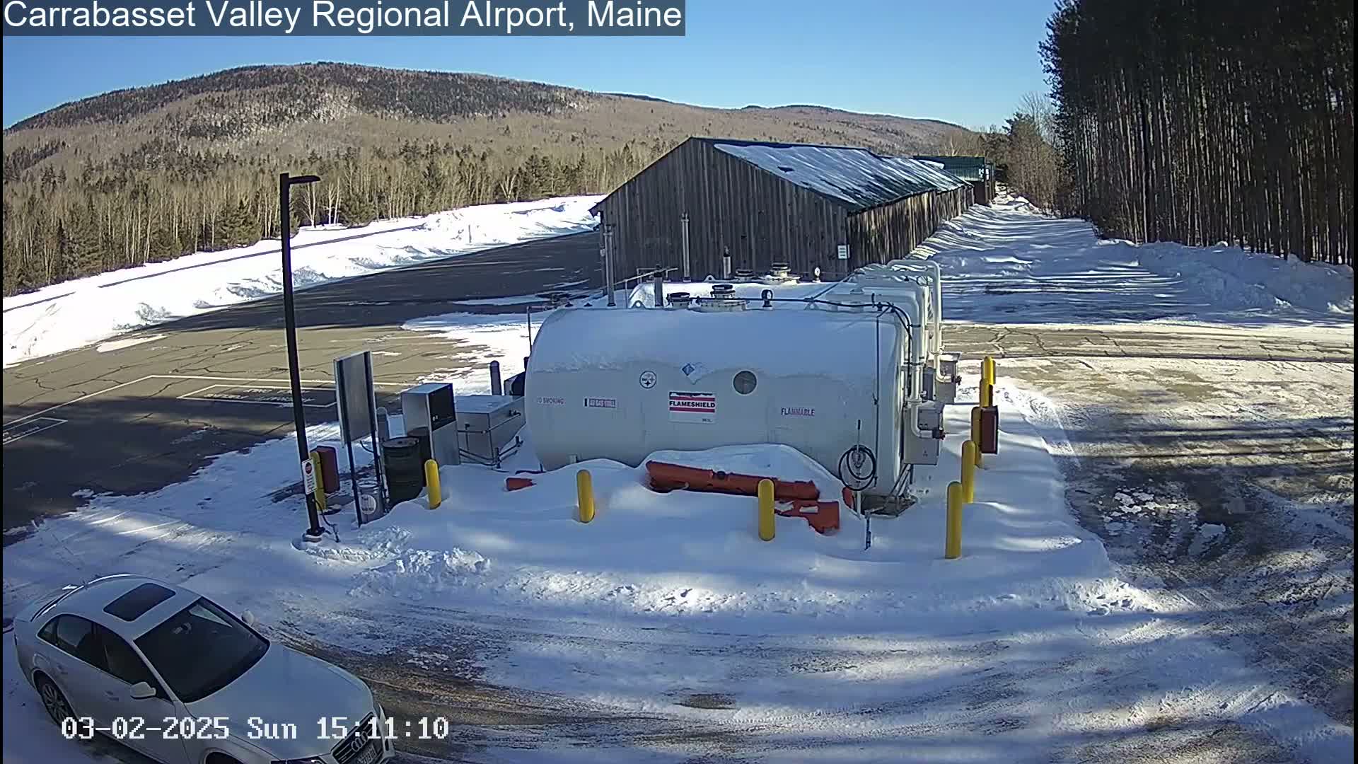 A snowy airport parking lot with a large white fuel tank, a nearby building, and a silver car are visible under a sunny sky.