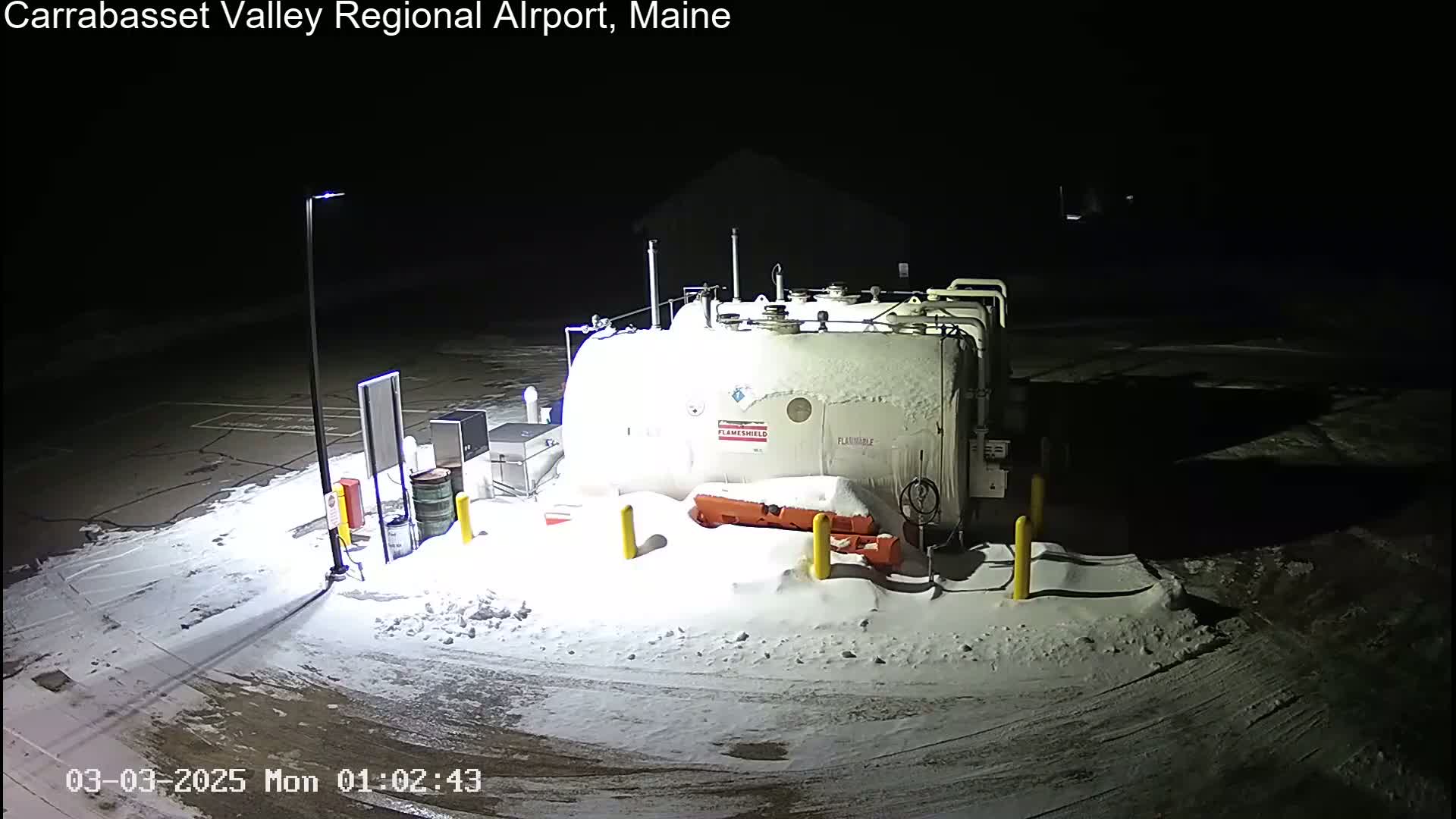 A large, snow-covered fuel tank sits in a snow-covered parking lot at night, illuminated by a single lamppost.