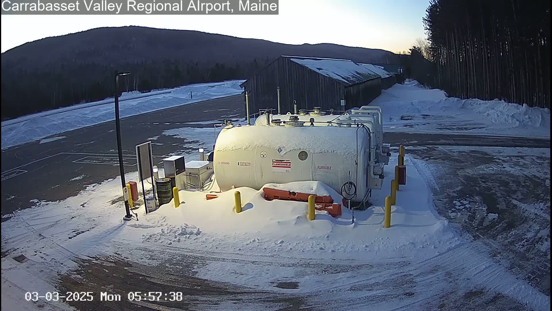Several large, snow-covered fuel tanks sit in a snow-covered parking lot next to a dark-colored building, with a mountain in the background under a twilight sky.