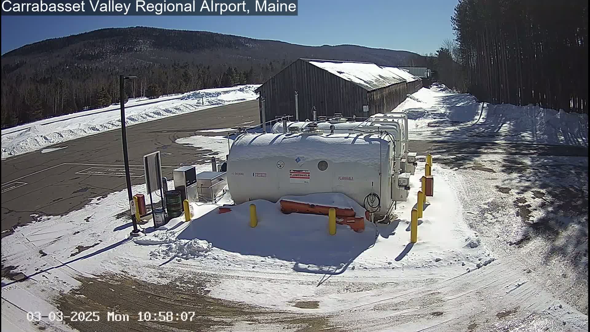 A snow-covered fuel storage area with several large tanks sits in front of a long wooden building on a sunny day, with mountains visible in the background.