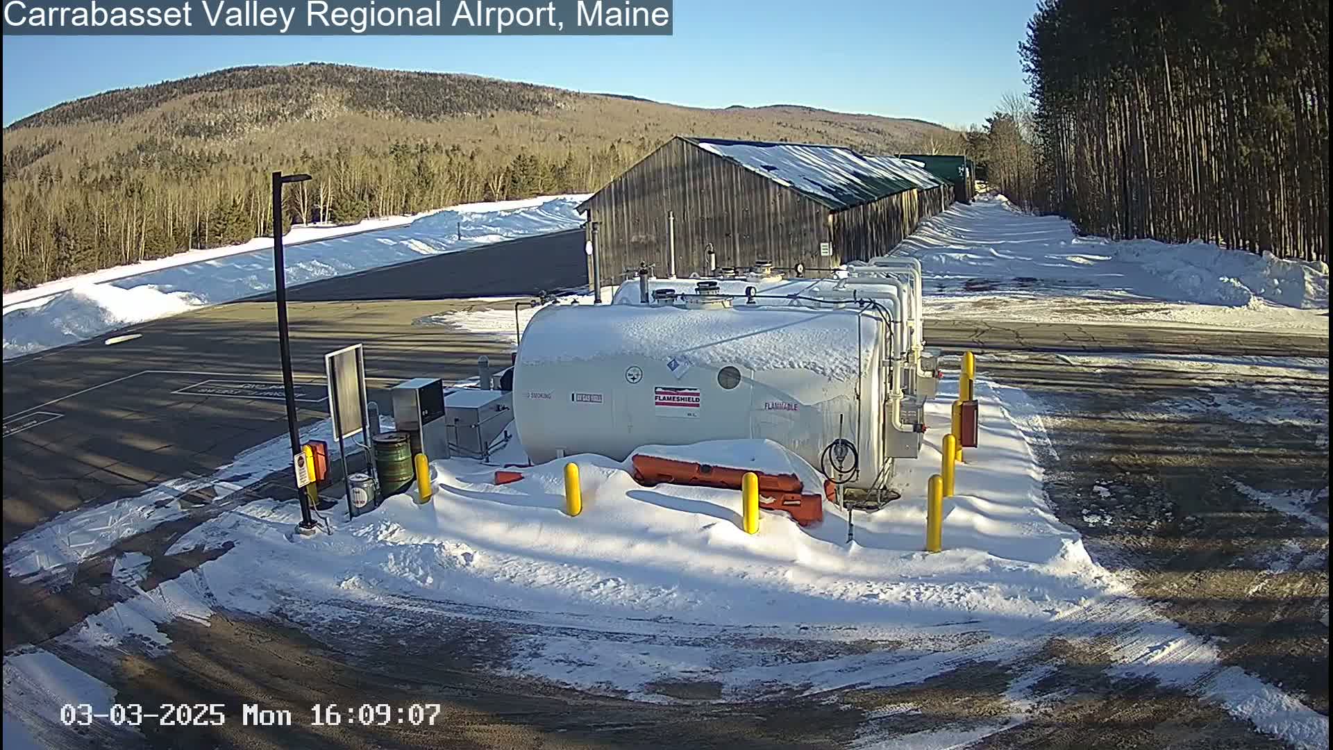 A snow-covered airport fuel depot is situated near a long, snow-dusted building, under a sunny winter sky.