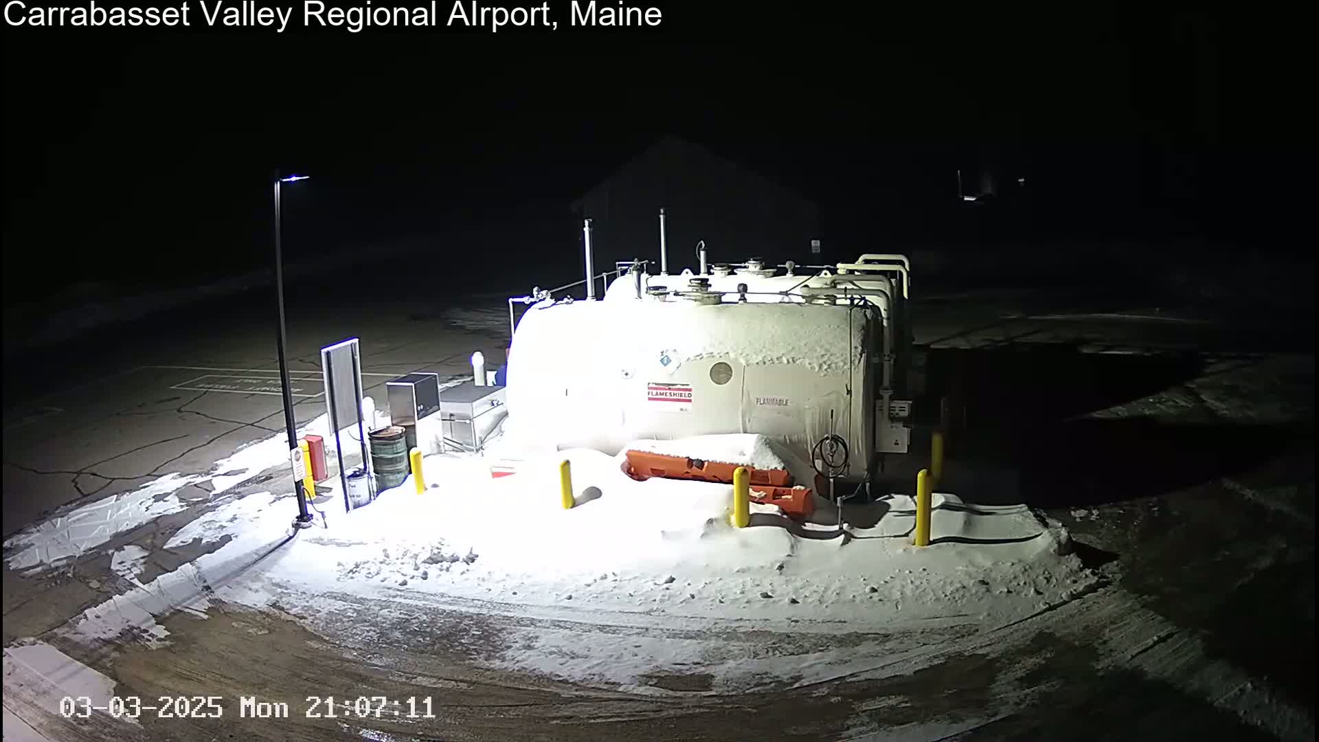 A large, snow-covered fuel tank sits in a snow-covered parking lot at night, illuminated by a single lamppost.