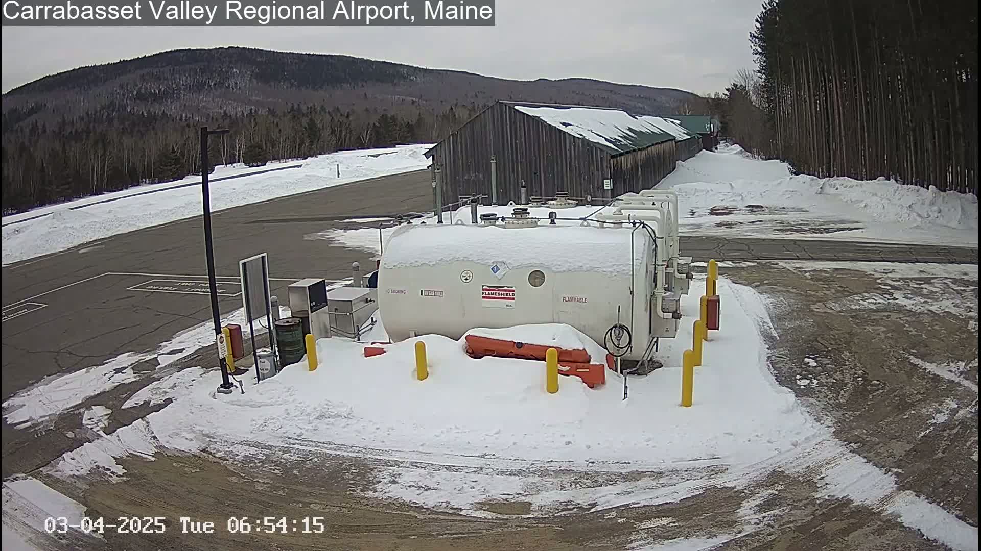 A snow-covered fuel storage tank sits in a snowy airfield parking lot next to a large wooden building, with a mountain range visible in the background under a cloudy sky.