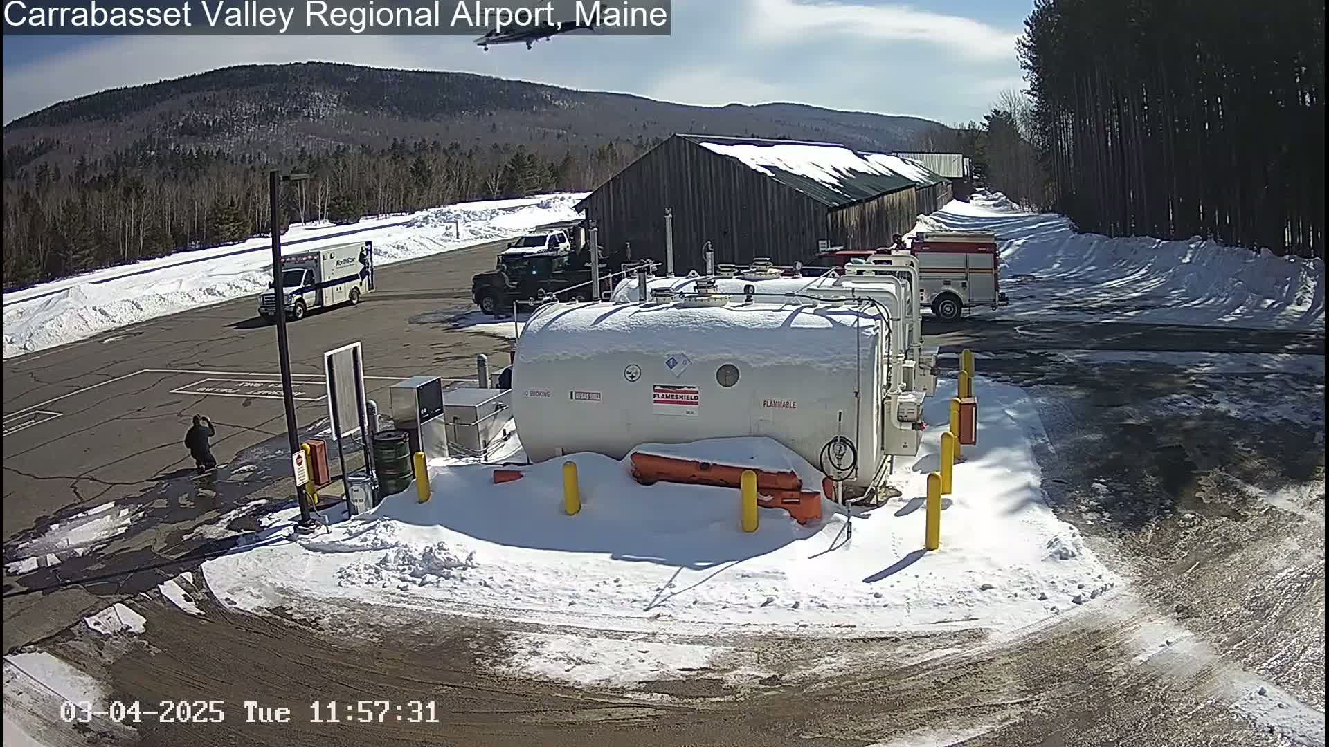A snowy airport tarmac contains several vehicles, including an ambulance, a pickup truck, and a firetruck, near large fuel tanks and a barn under a sunny sky.
