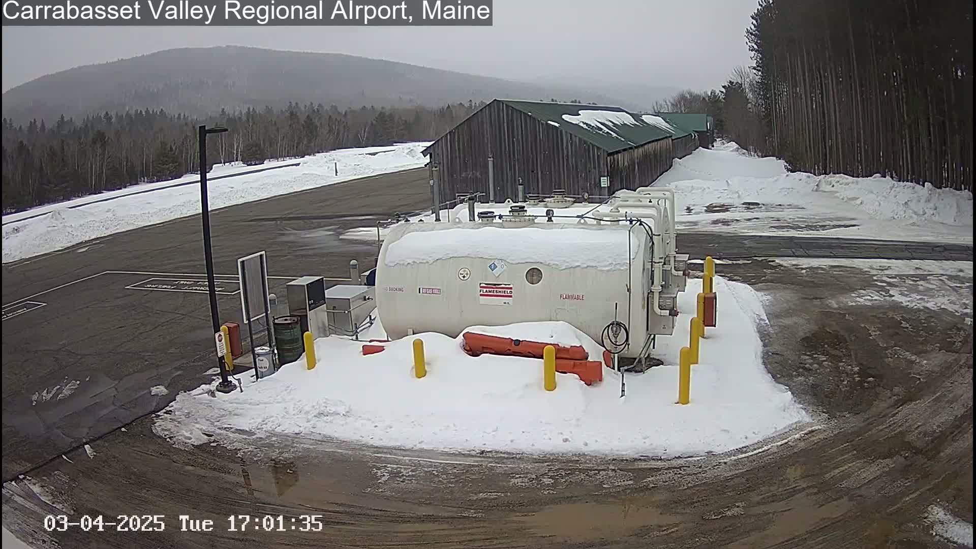 A snow-covered airport fuel storage tank sits in a parking lot next to a large wooden building, with a background of snow-covered trees and a hazy mountain range under an overcast sky.
