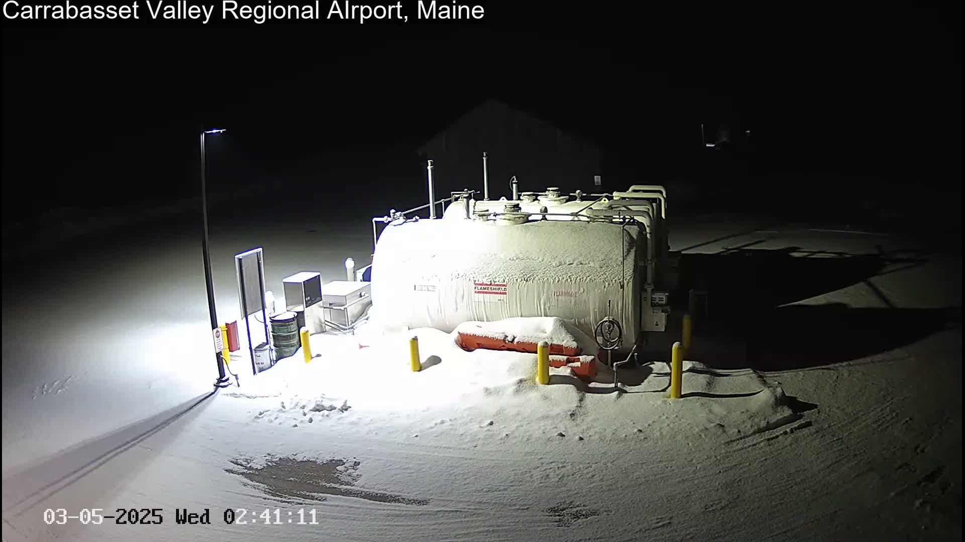 A large, snow-covered fuel tank sits in a snow-covered area at night, illuminated by a single lamppost.