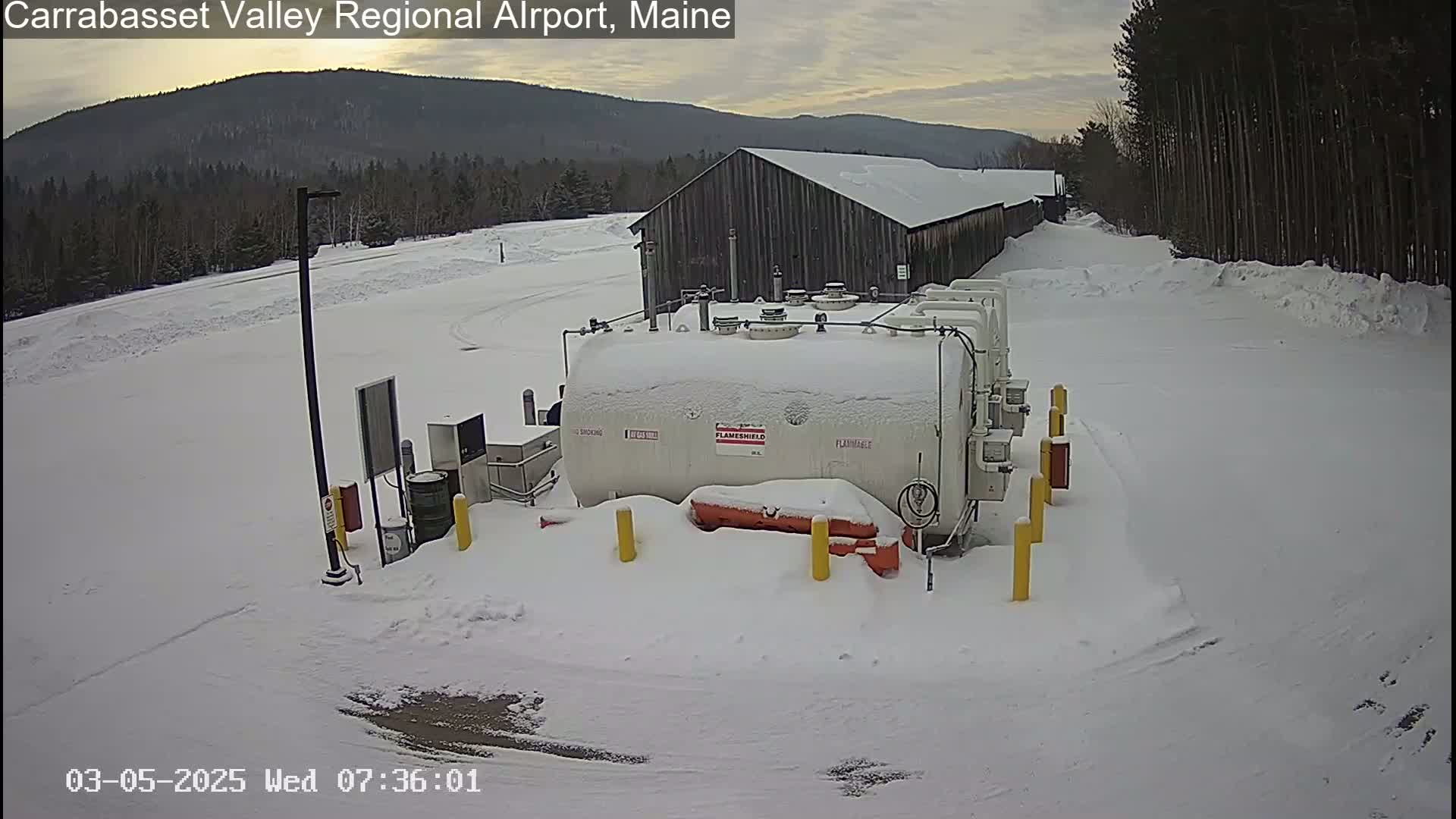 A snow-covered fuel tank sits in front of a snow-covered building near a wooded area under an overcast sky.