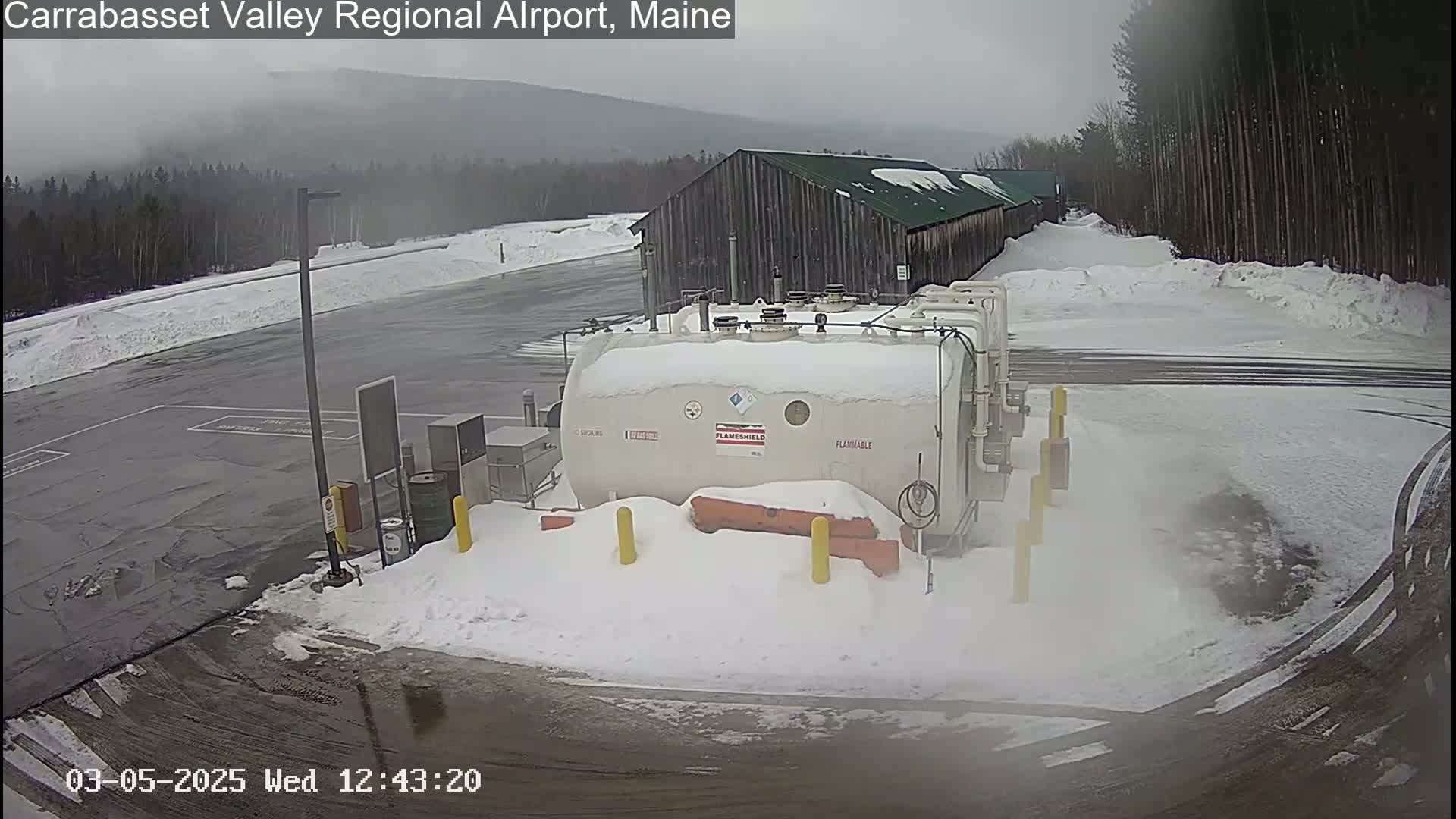 A snow-covered airport tarmac with a large, snow-covered fuel tank near a wooden building under an overcast sky.