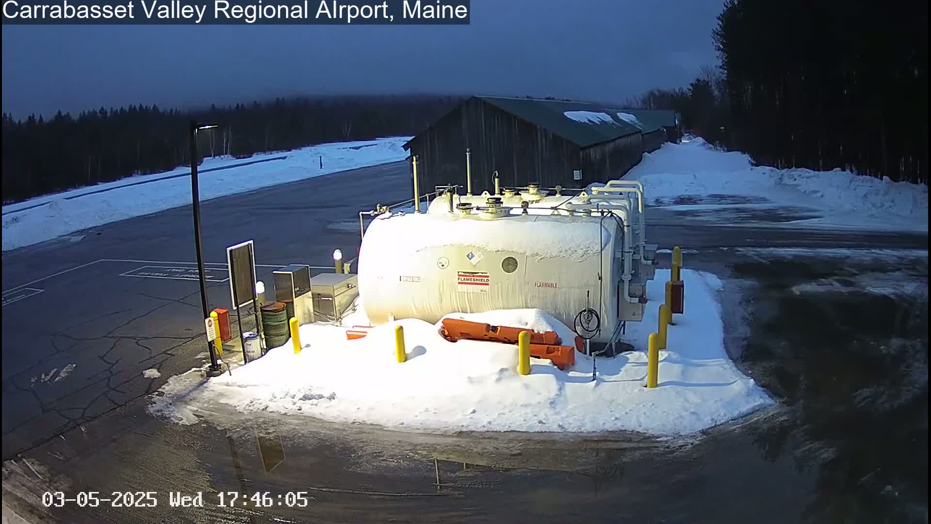 A large, snow-covered fuel tank sits in a snow-covered parking lot next to a dark-colored building, under a twilight sky.