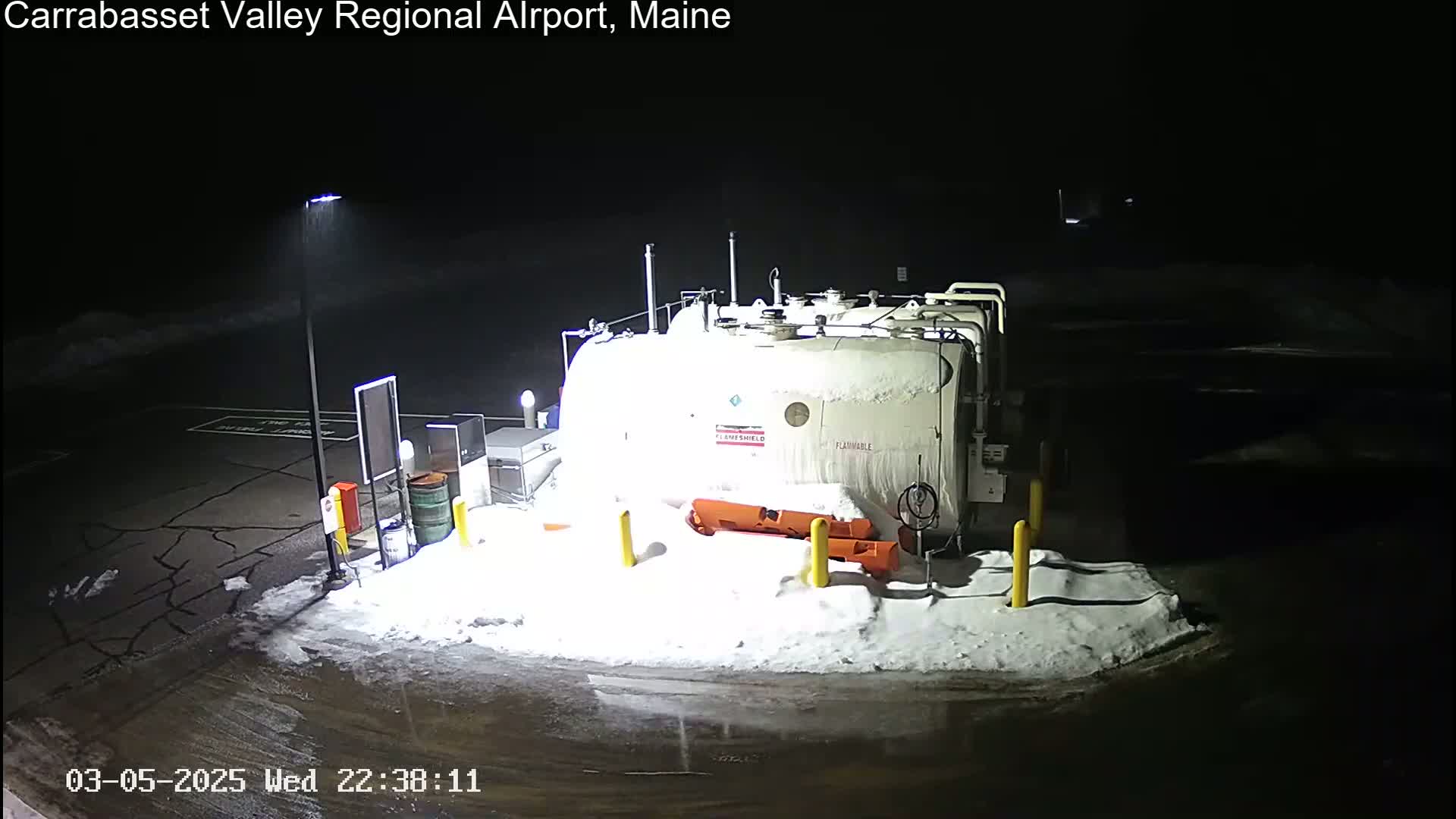 A large, snow-covered fuel tank sits in a snow-covered parking lot at night, illuminated by a single overhead lamp.
