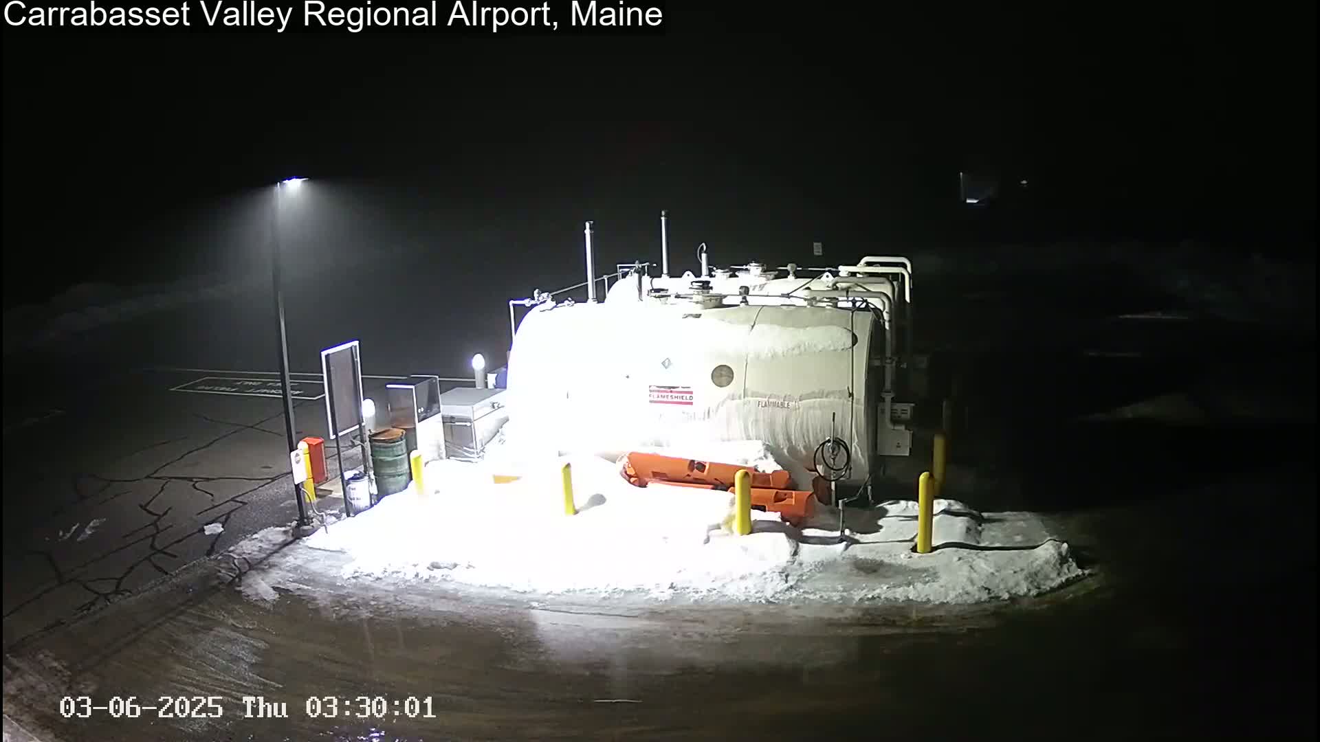 A large, snow-covered fuel tank sits in a snow-covered area at night, illuminated by a nearby light post.