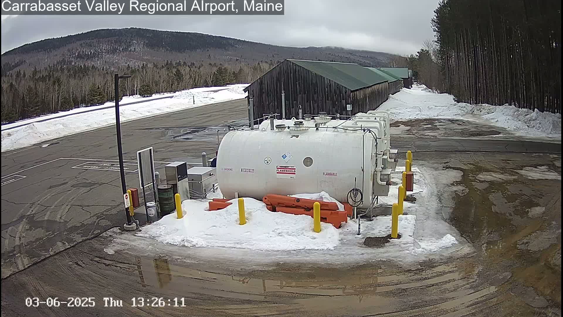 A large white fuel tank sits in a snowy airfield parking lot near a dark-colored building, under an overcast sky.