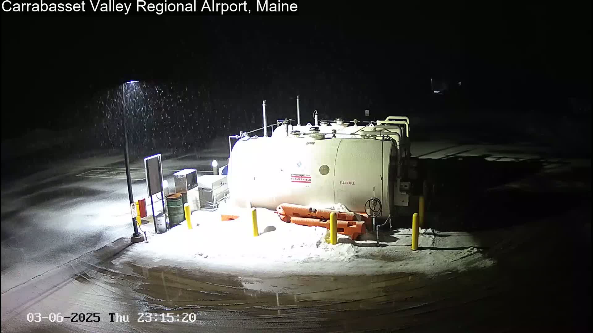 A large, white fuel tank sits in a snow-covered airport parking lot during a light snowfall at night.