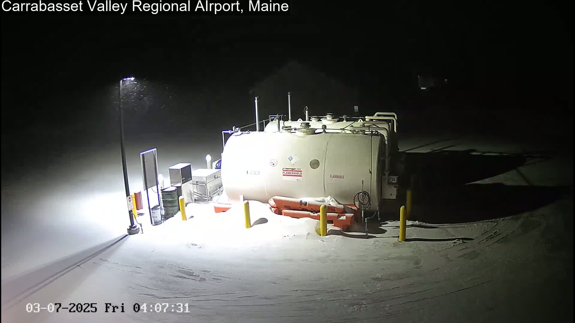 A large, white, cylindrical fuel tank sits in a snow-covered area at night under a light pole during a light snowfall.