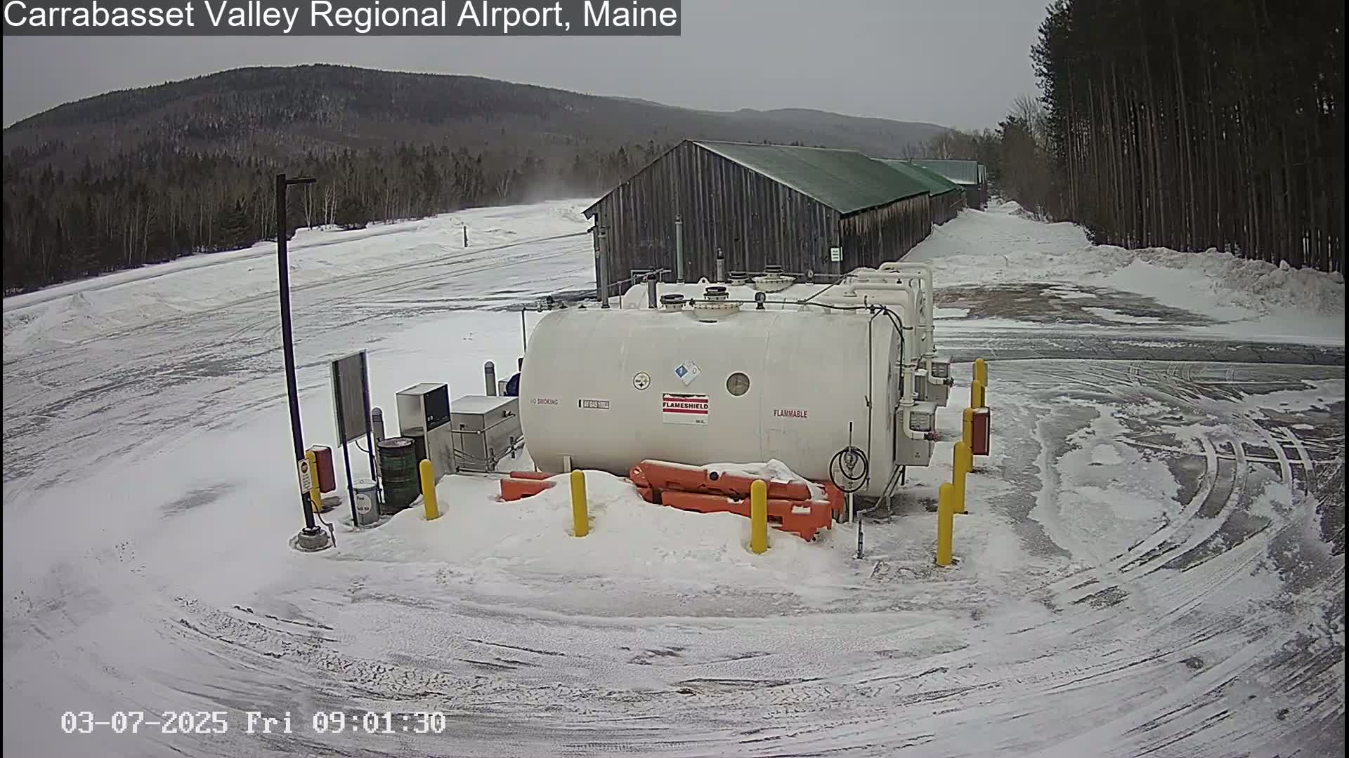 A large white fuel tank sits in a snowy airport parking lot next to a long wooden building, with a background of snow-covered trees and mountains under a cloudy sky.