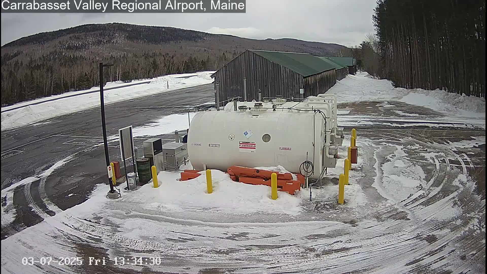 A large white fuel tank sits in a snow-covered parking lot near a long, dark-colored building, with a background of snow-covered hills and trees under a cloudy sky.