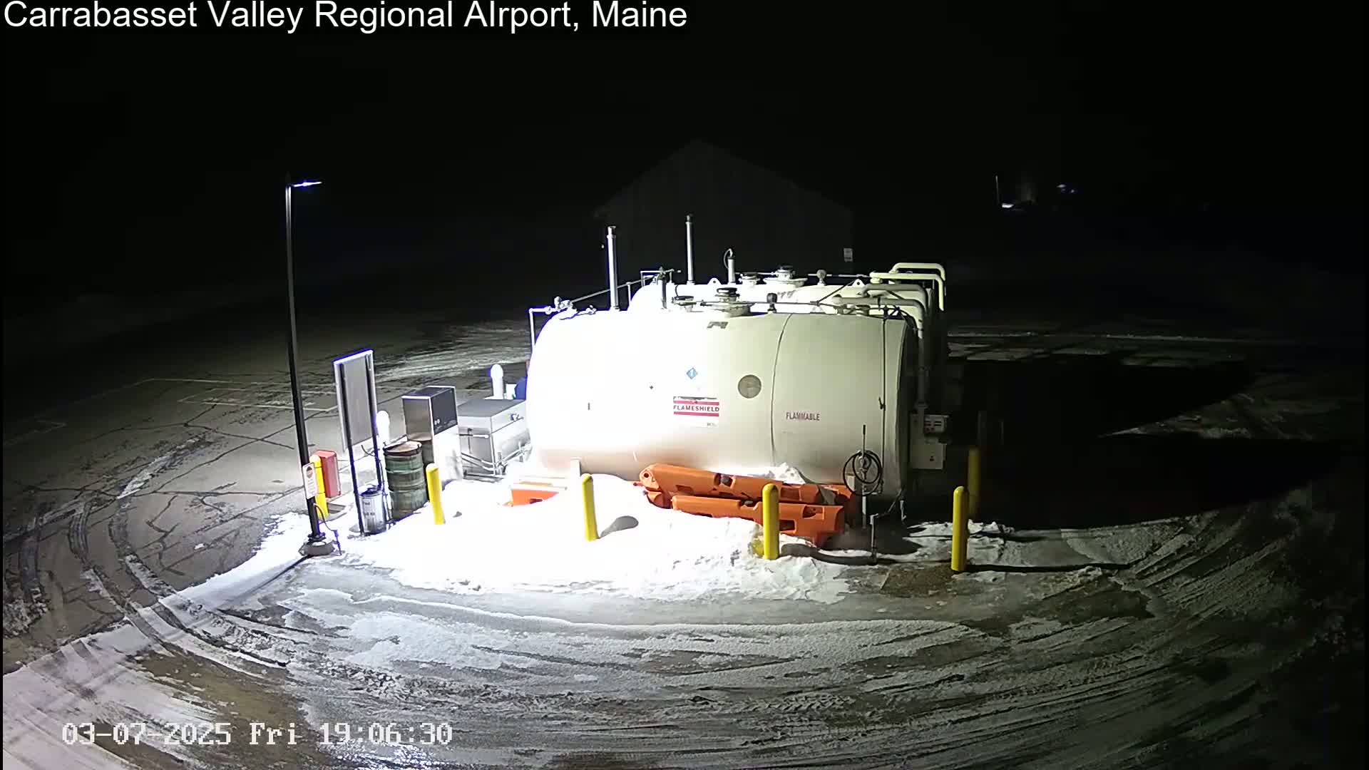 A large, white fuel tank sits in a snow-covered parking lot at night, illuminated by a single lamppost.