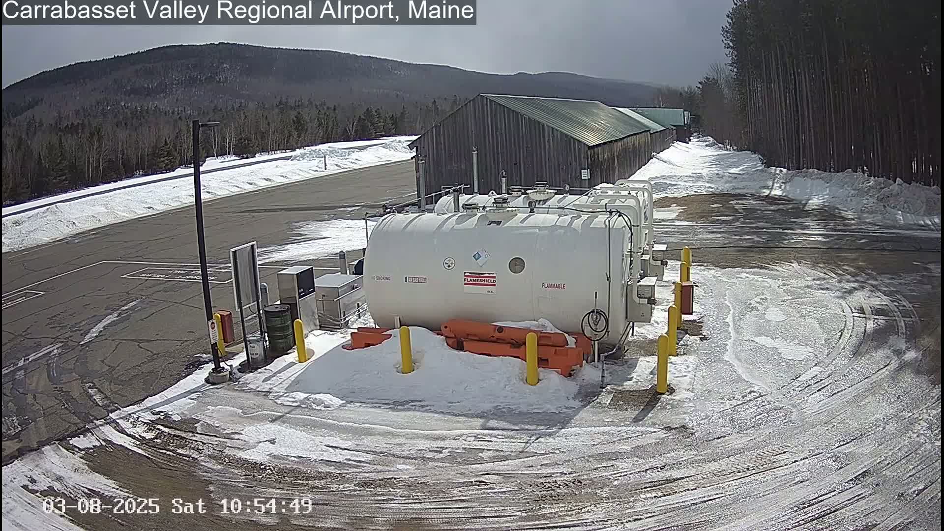 A snowy airport tarmac with several large white tanks and a long, low building in the background under a partly sunny sky.