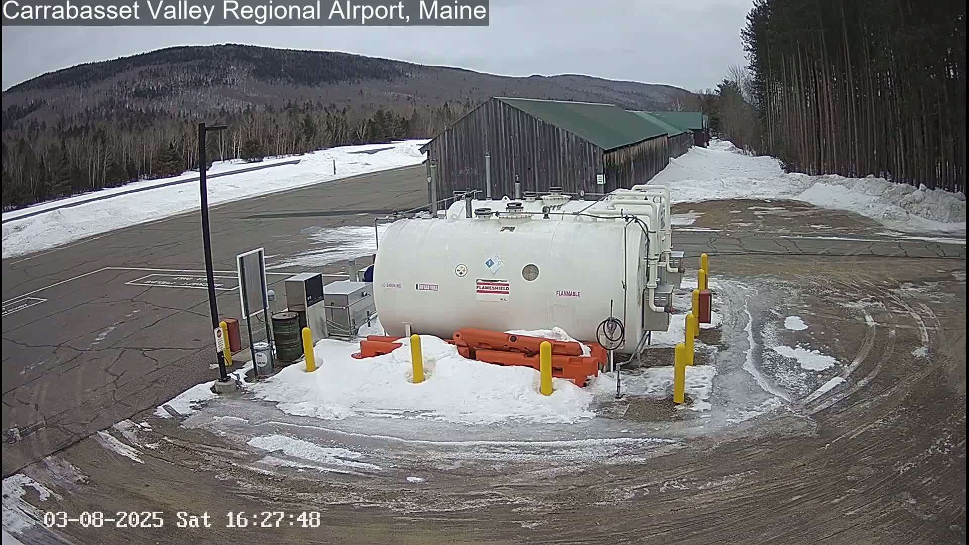 A snowy airfield with several large white fuel tanks and a long, dark-colored building in the background under an overcast sky.