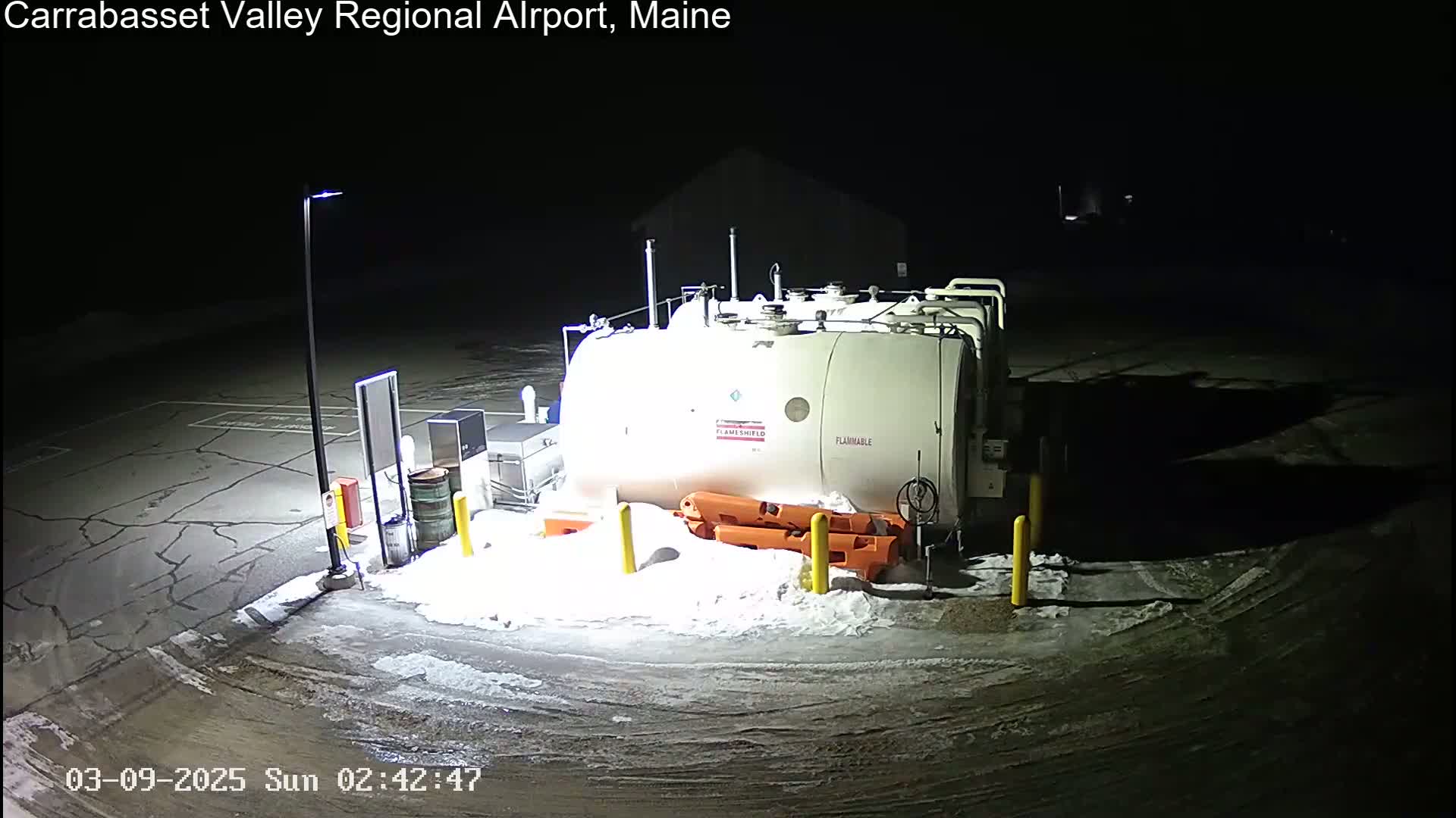 A large, white, flammable tank sits in a snow-covered parking lot at night, illuminated by a single light pole.