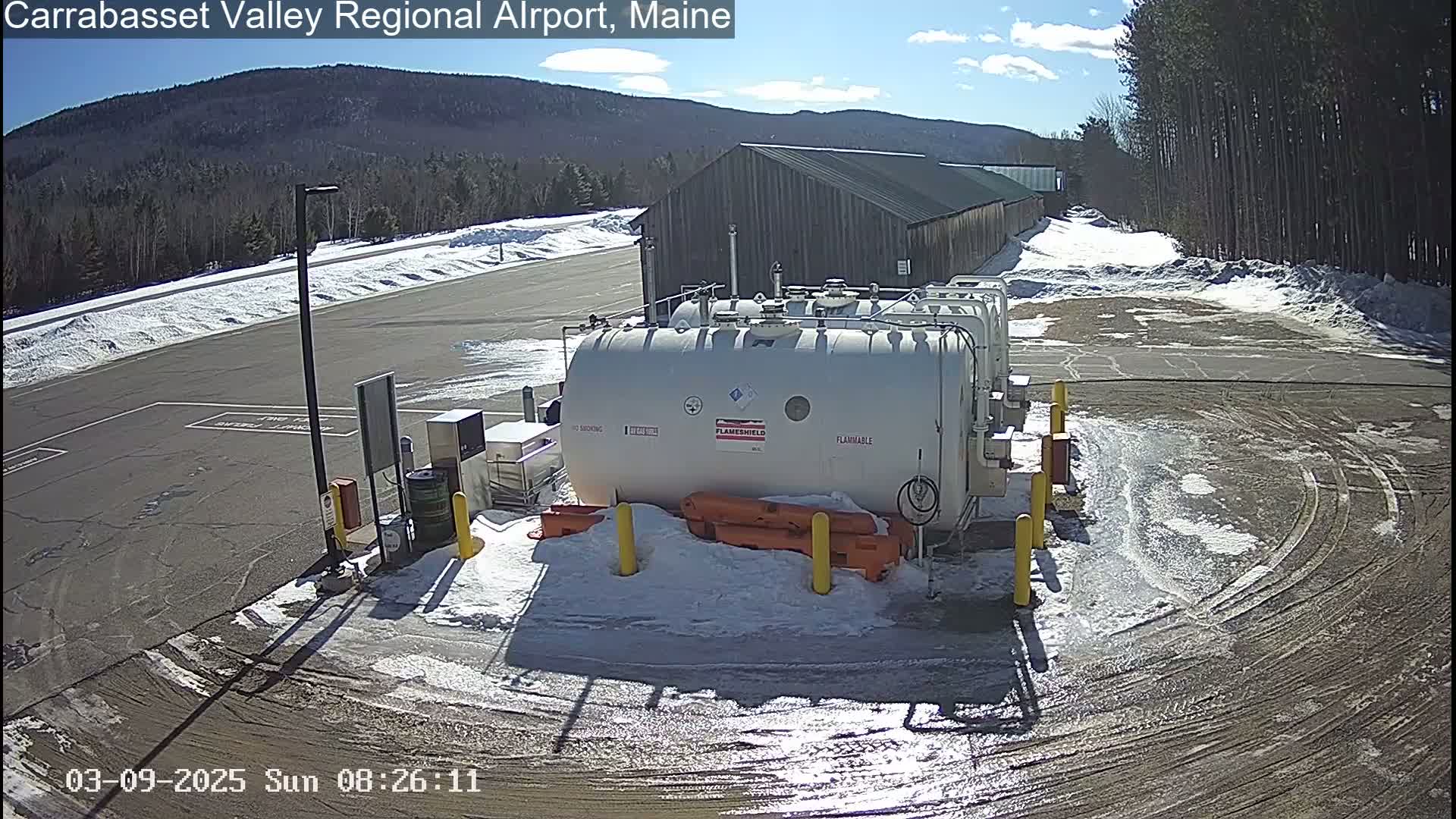 A large white fuel tank sits in a snow-covered airport parking lot under a sunny sky, with a long, low building and distant mountains visible in the background.