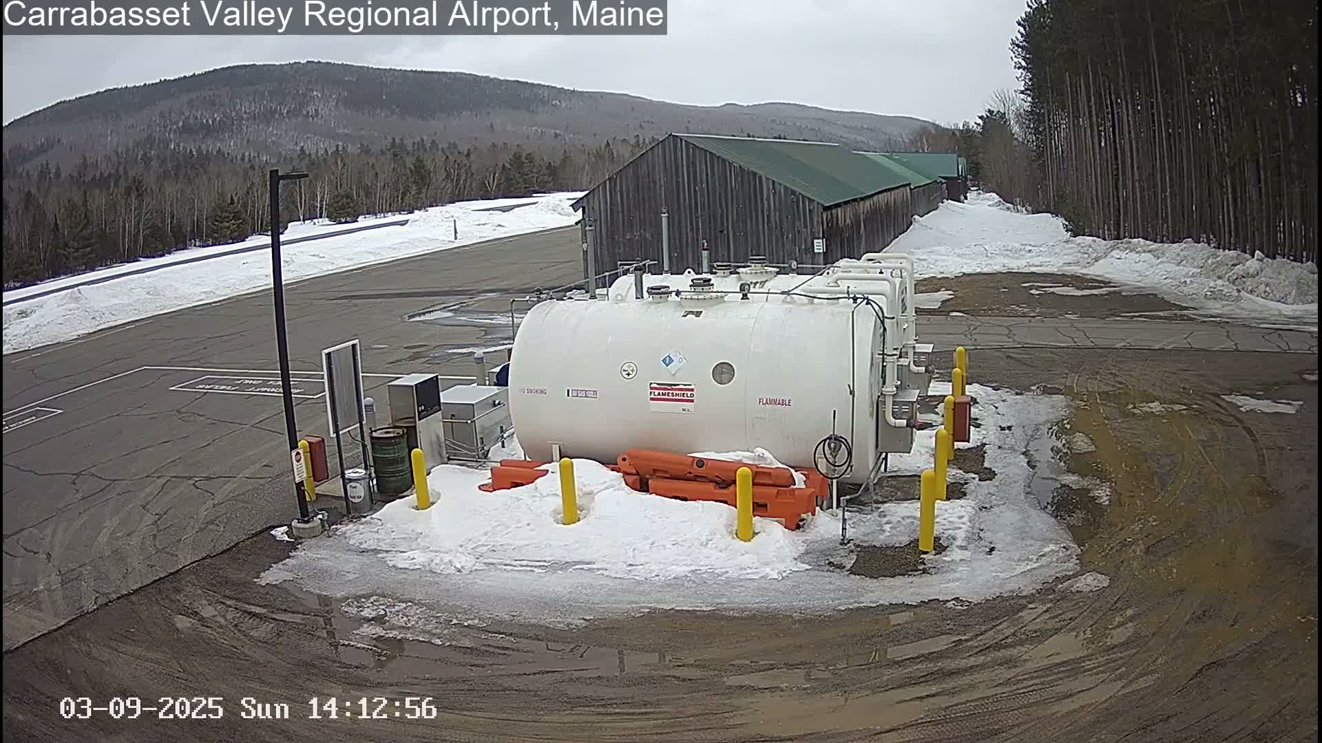 A large white fuel tank sits in a snowy, muddy parking lot near a wooded area and a long building under an overcast sky.