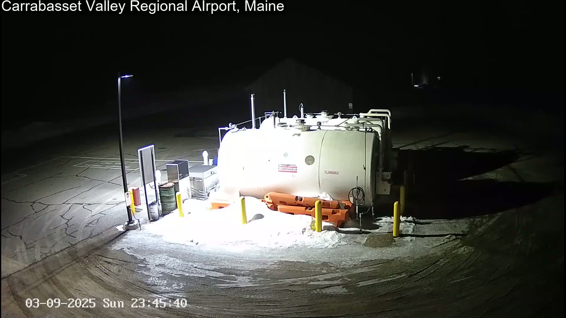 A large, cylindrical fuel tank sits in a snow-covered parking lot at night, illuminated by a single lamppost.