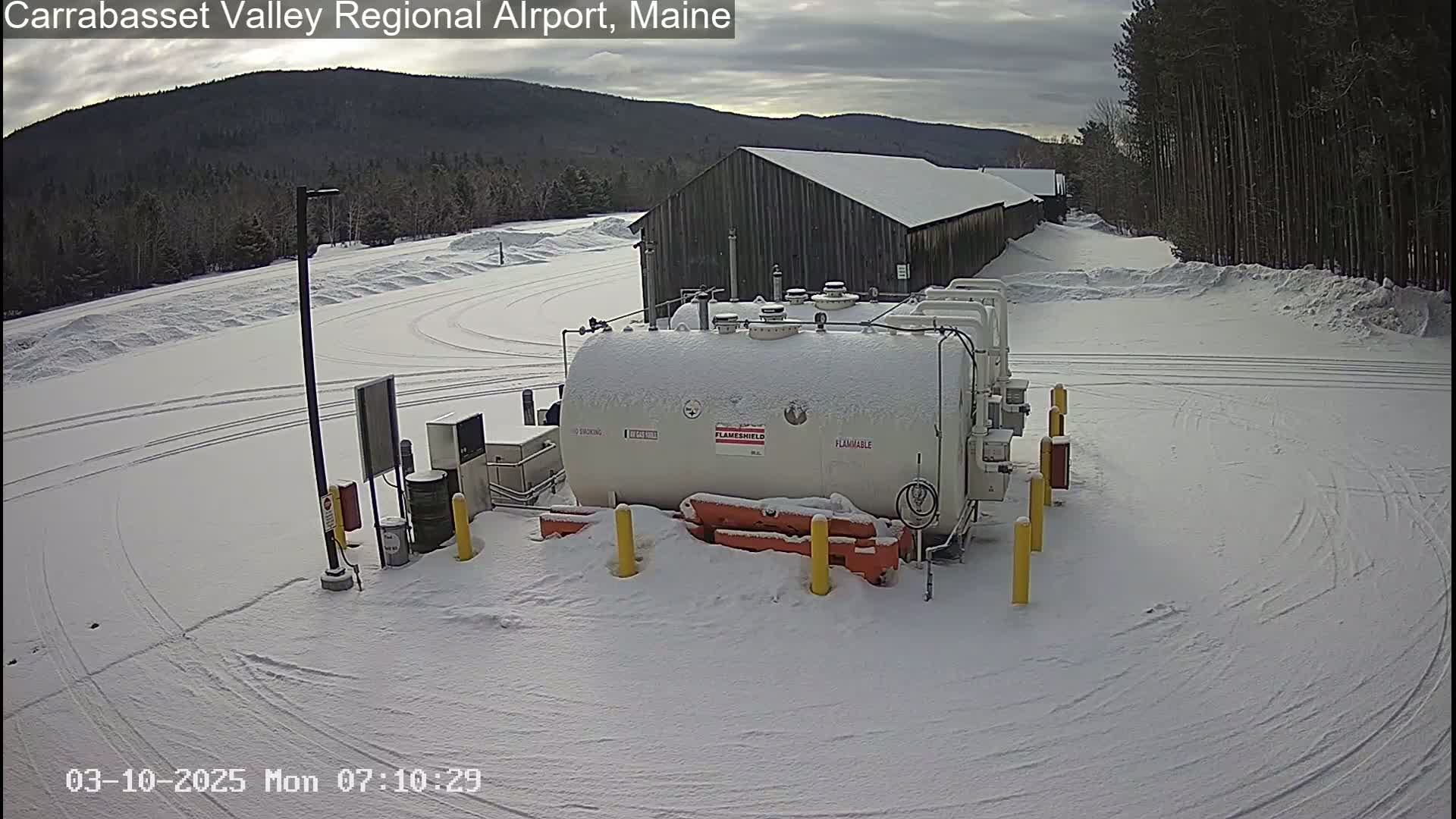 A snow-covered airfield shows a large white tank, marked "flammable," near a long gray building, with tire tracks in the snow and a cloudy sky.
