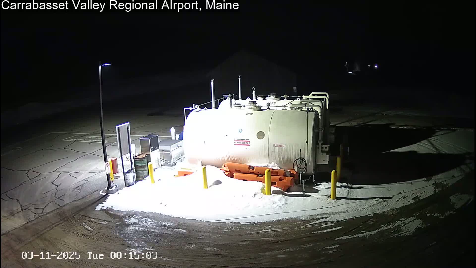 A large, white fuel tank sits in a snow-covered airport parking lot at night, illuminated by a single lamppost.