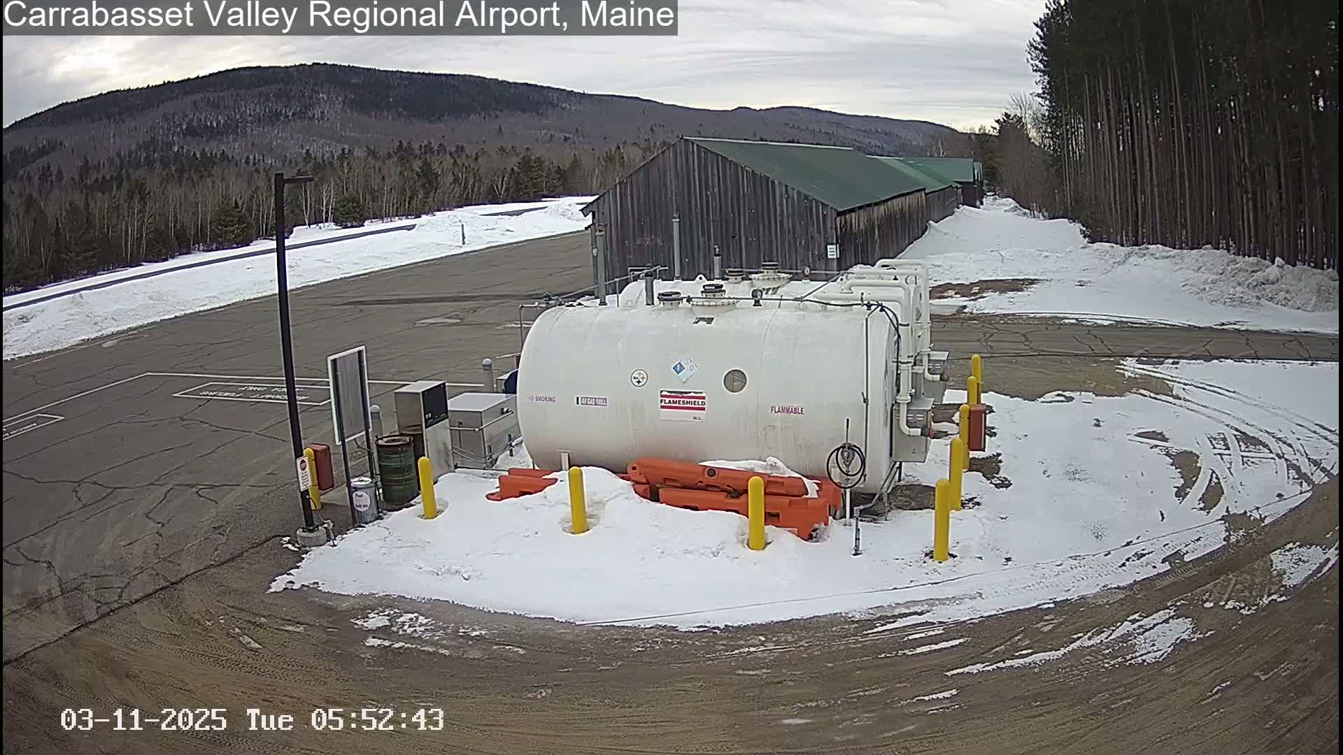 A large white fuel tank sits in a snow-covered airport parking lot next to a long, dark-colored building, with a background of a snow-covered mountain range under a cloudy sky.