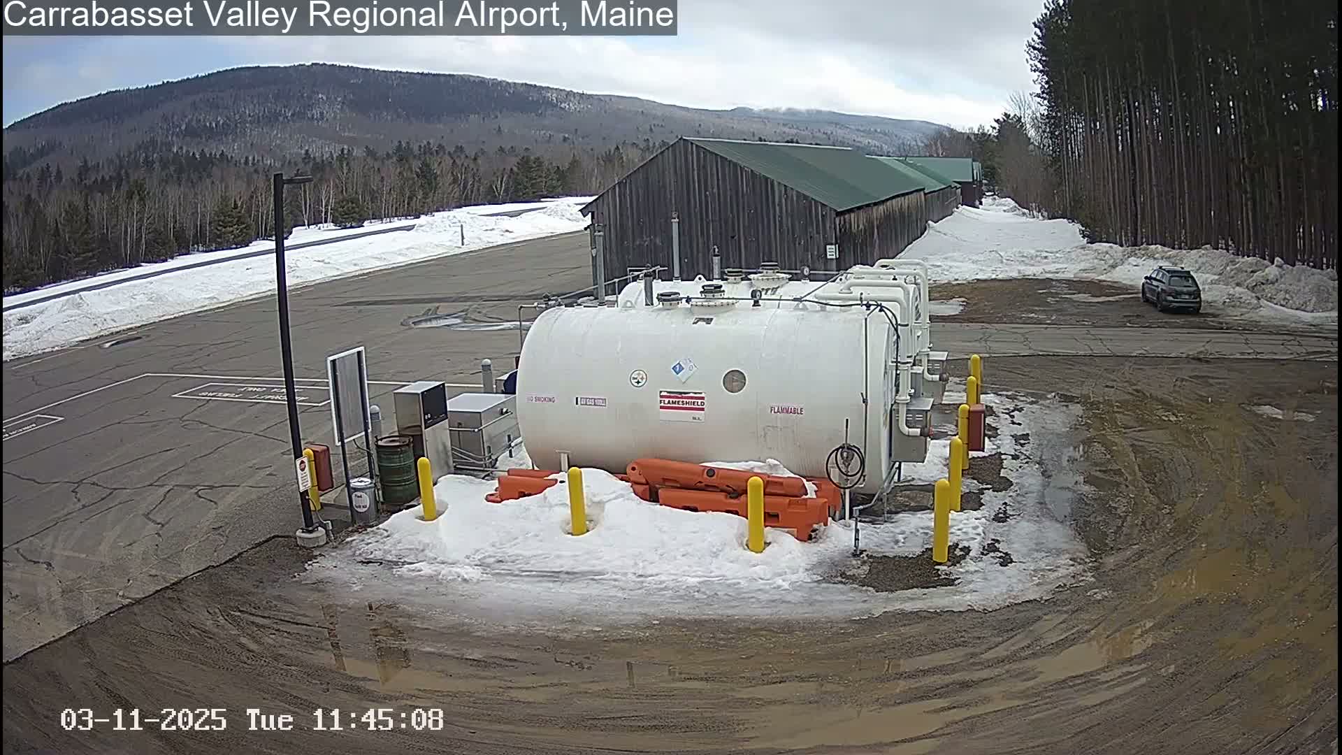 A large white fuel tank sits in a snowy, muddy parking lot near a long building, with a car parked in the distance under an overcast sky.