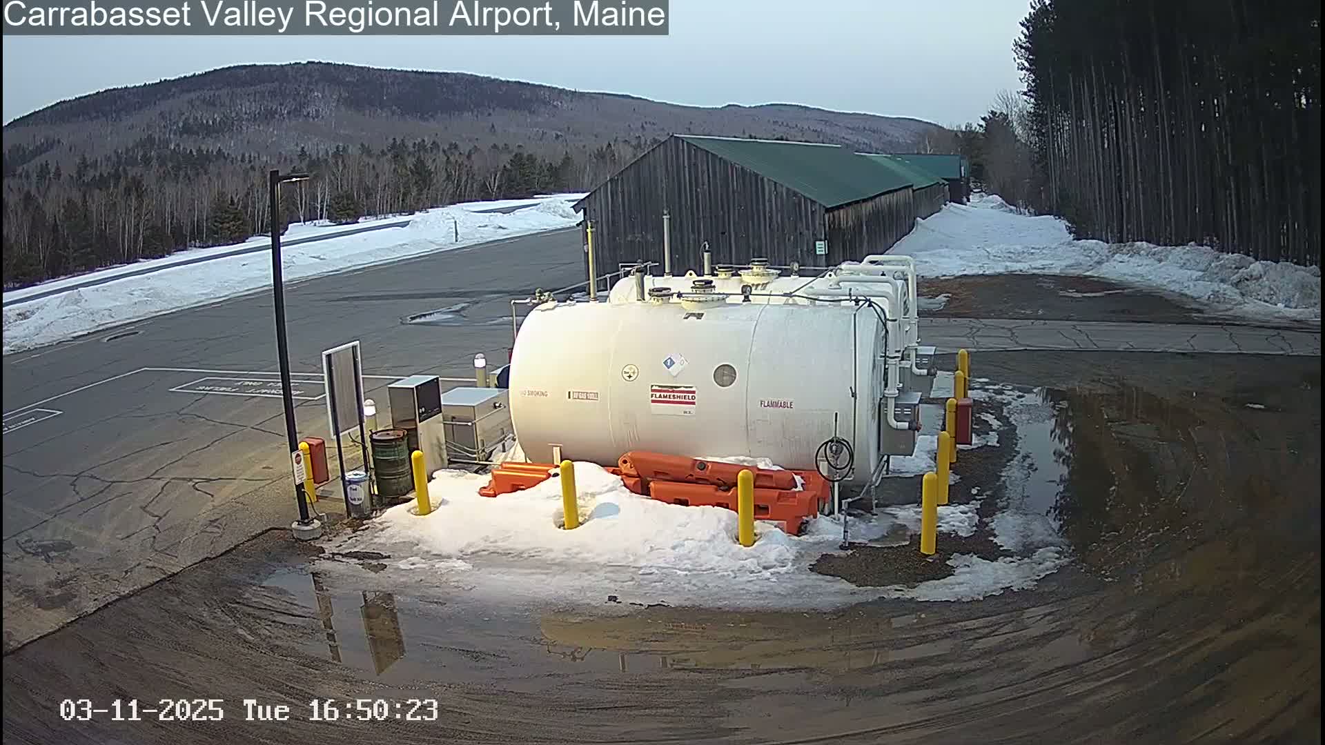 A large white fuel tank sits in a snowy, partially paved area next to a building, with a background of a wooded hillside under a cloudy sky.
