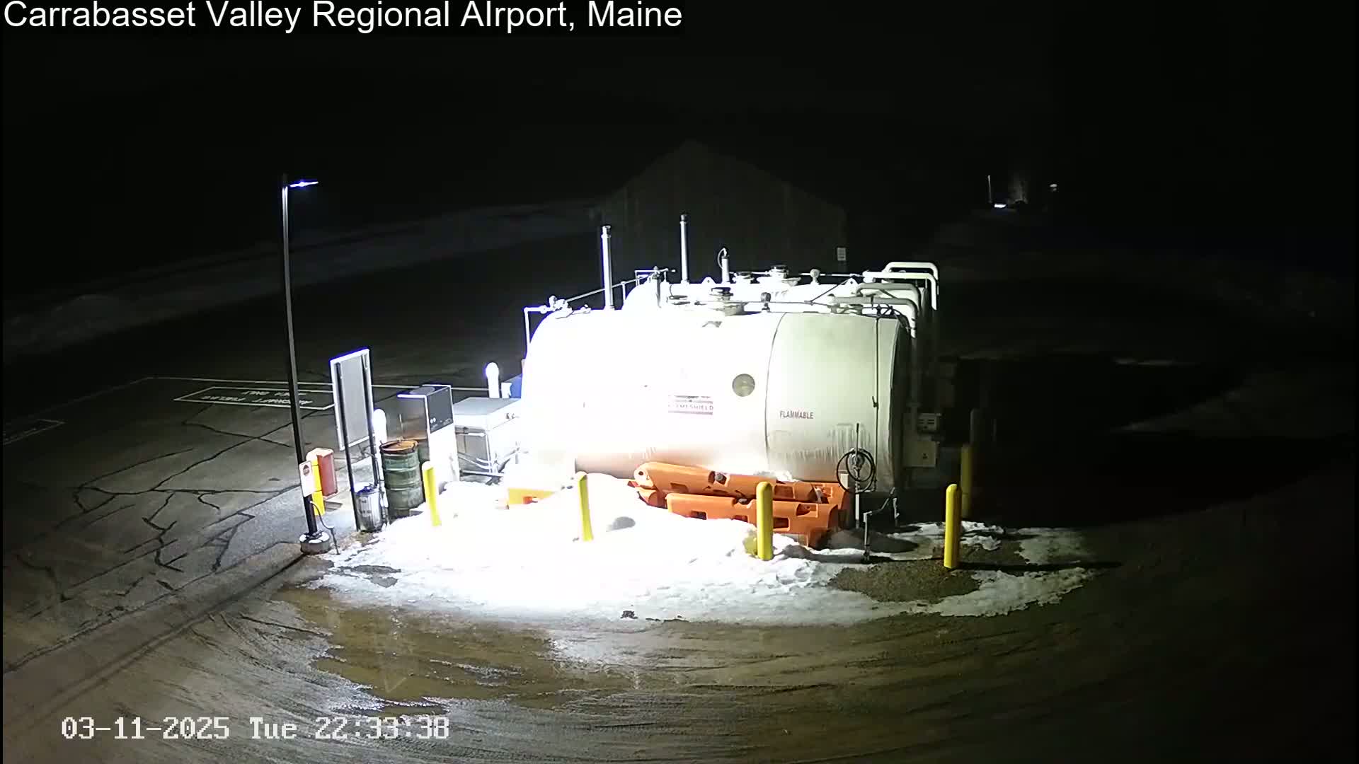 A large, white fuel tank sits in a snow-dusted parking lot at night, illuminated by a single lamppost.
